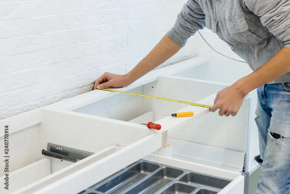 Person measuring a white cabinet frame with a tape measure in a workshop.