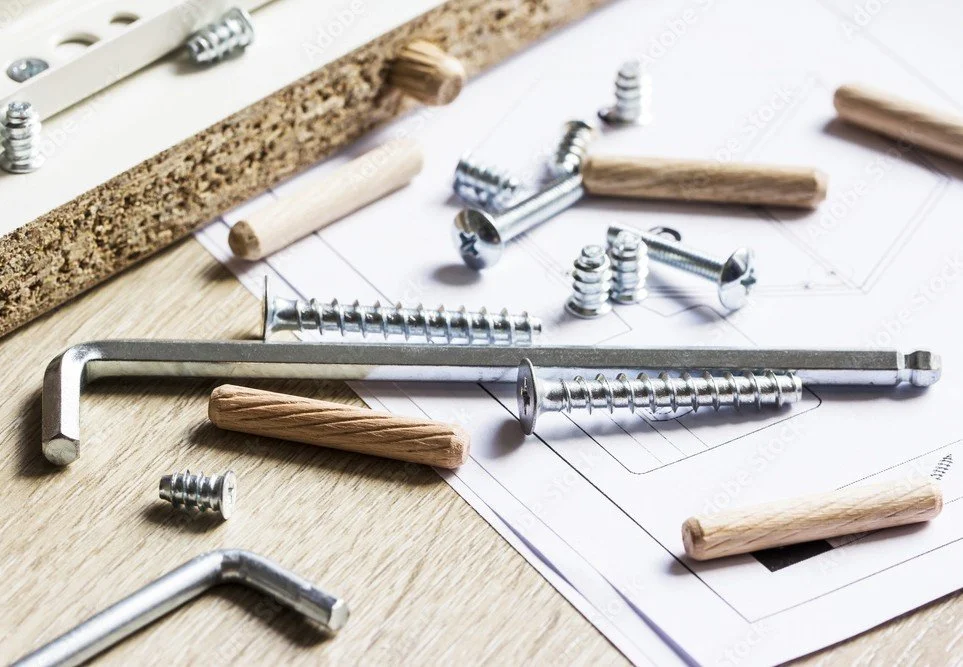 Construction tools including screws, bolts, an Allen wrench, and wooden dowels on a work surface with blueprints.