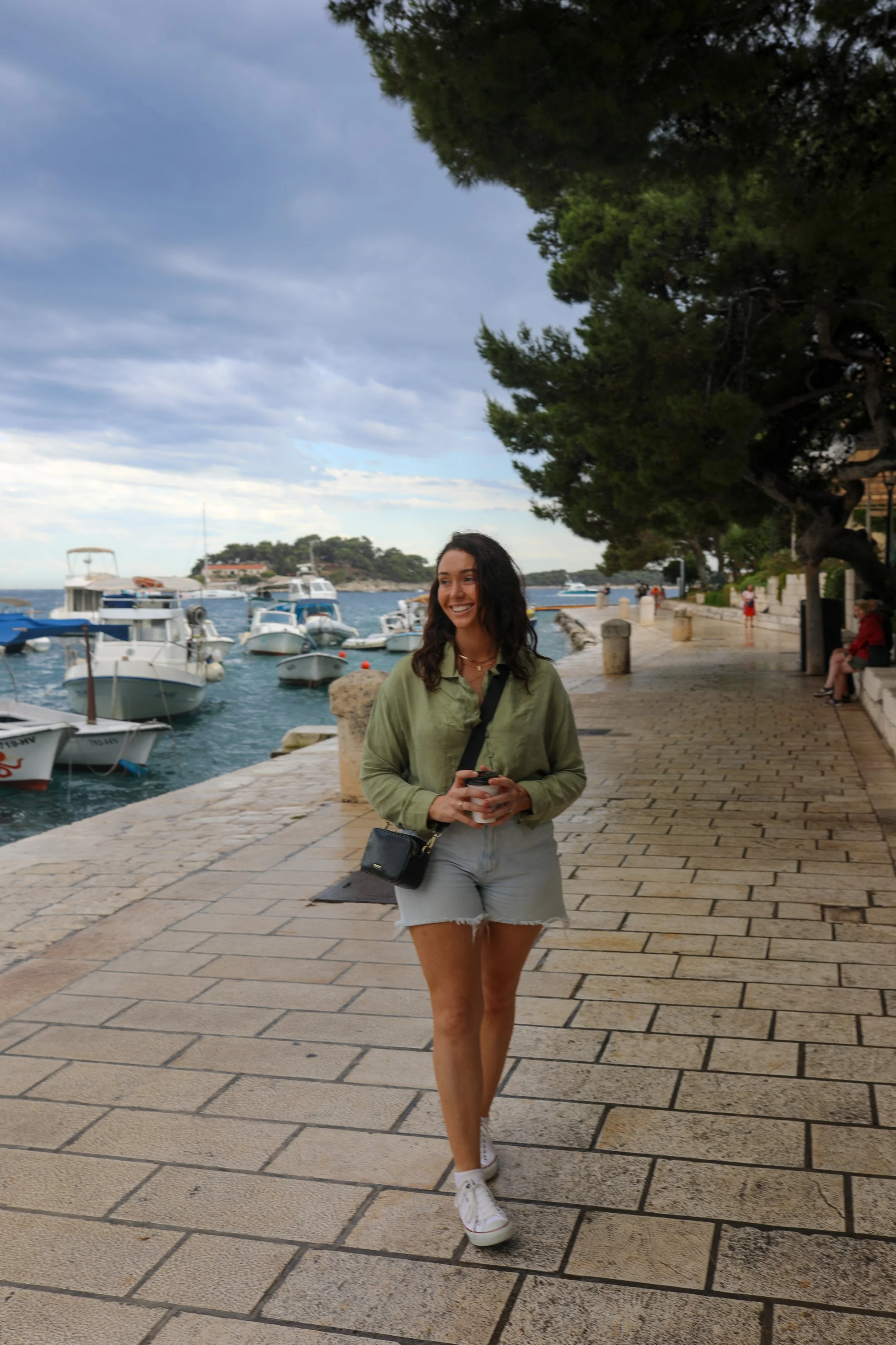 A woman walking along a harbor promenade with boats docked nearby, wearing a green jacket, gray shorts, white sneakers, smiling and holding a cup, trees and clouds in the background.