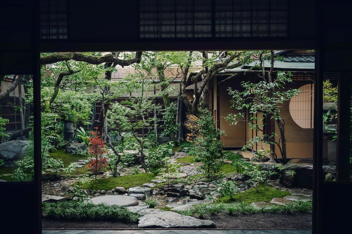 Traditional Japanese garden viewed through a shoji screen window, featuring lush greenery, small trees, rocks, and a stone pathway.
