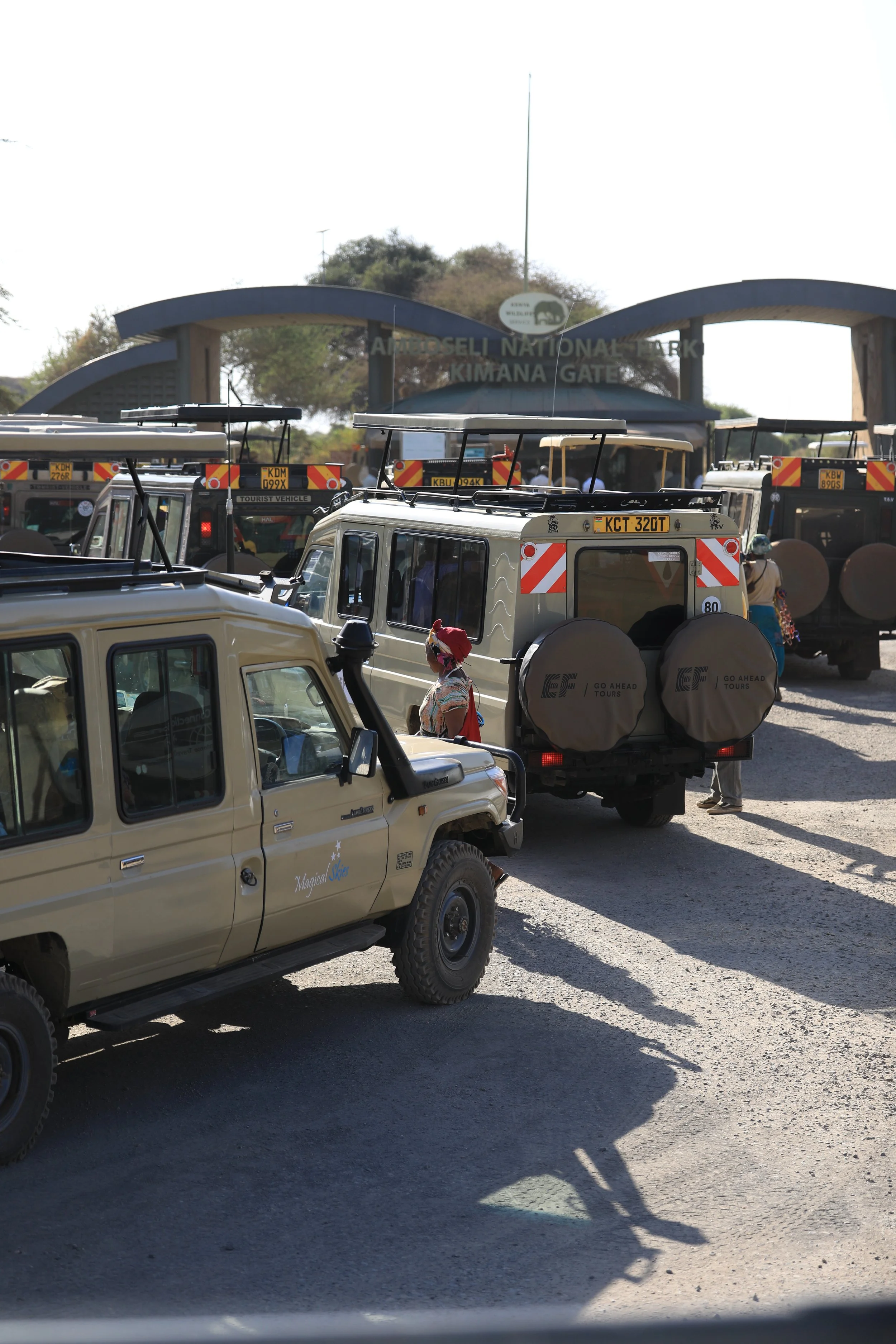 Tourist vehicles parked at the entrance of Amboseli National Park, Kenya, with a sign reading 'Kimana Gate' in the background and some people walking nearby.