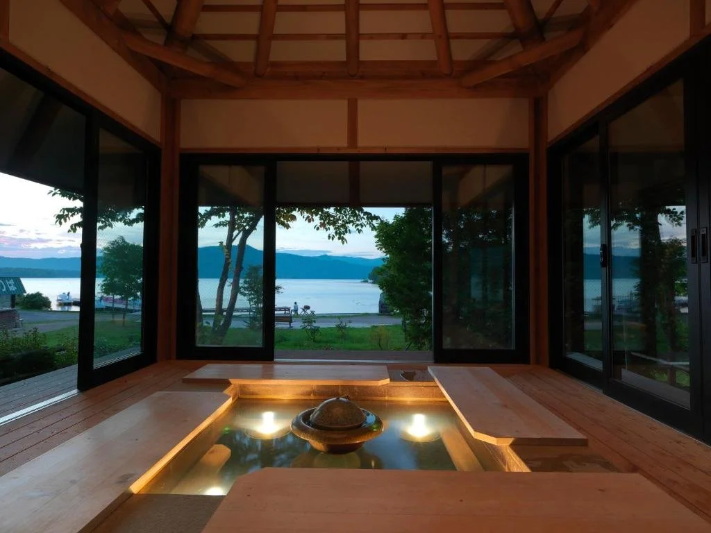Indoor hot spring bath with a view of the lake and mountains outside through large glass windows in a wooden structure.