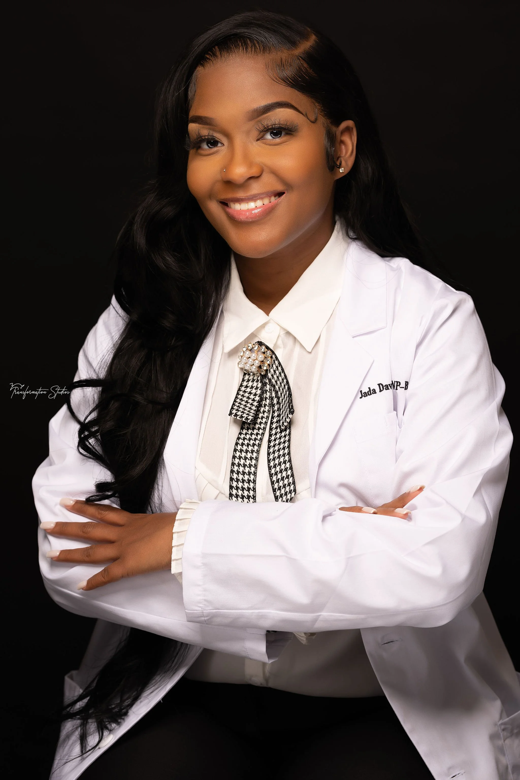 Professional woman wearing a white blazer with her arms crossed, smiling, with long black hair styled in waves, against a dark background.