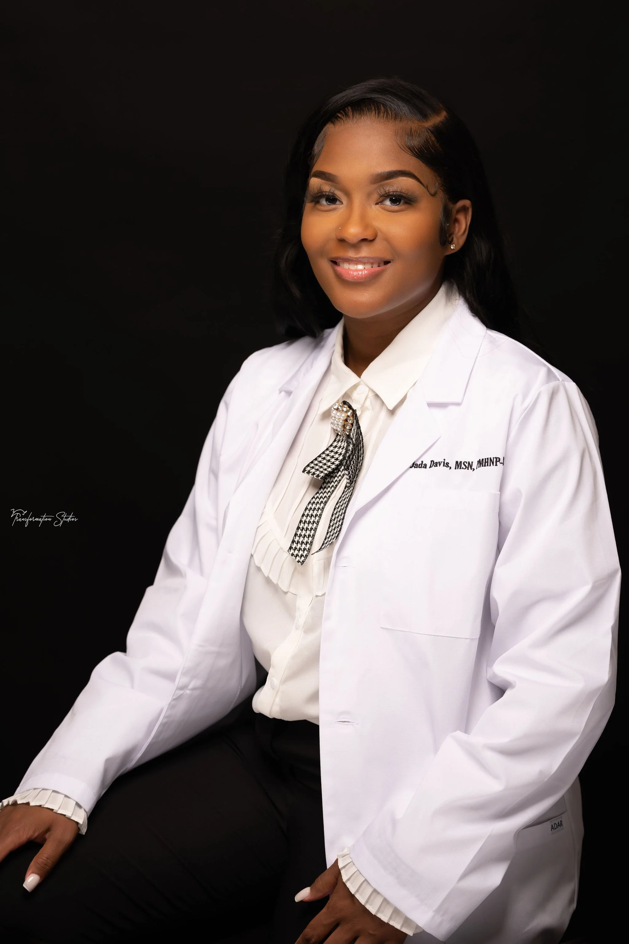 Professional woman in white lab coat smiling at the camera, seated against a black background.