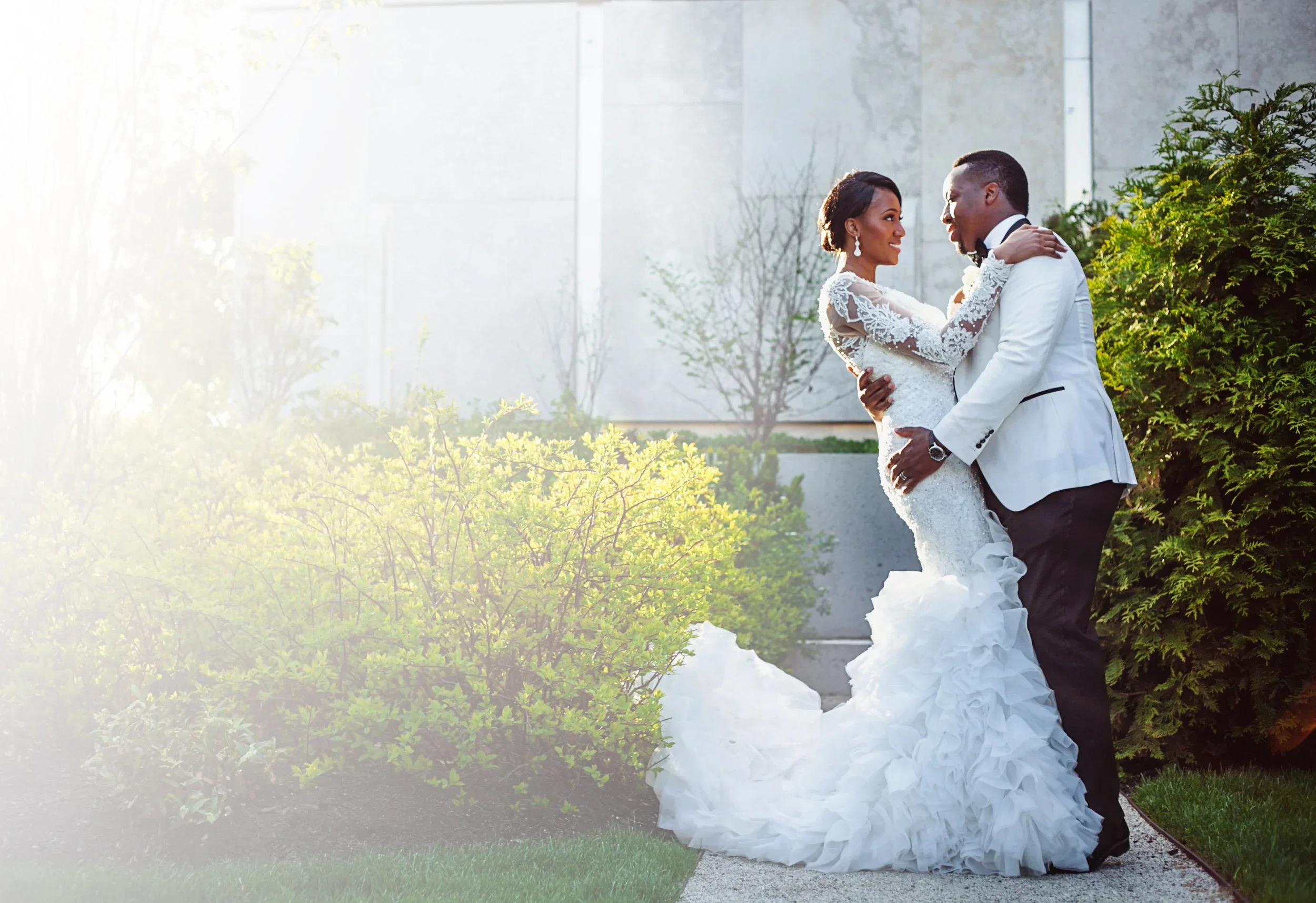 A wedding cake with a topper of a man and woman in wedding attire, standing close together, with the woman in a white wedding dress and the man in a black hat and suit. The cake is decorated with pink and white icing, roses, pearls, and horse shoes.