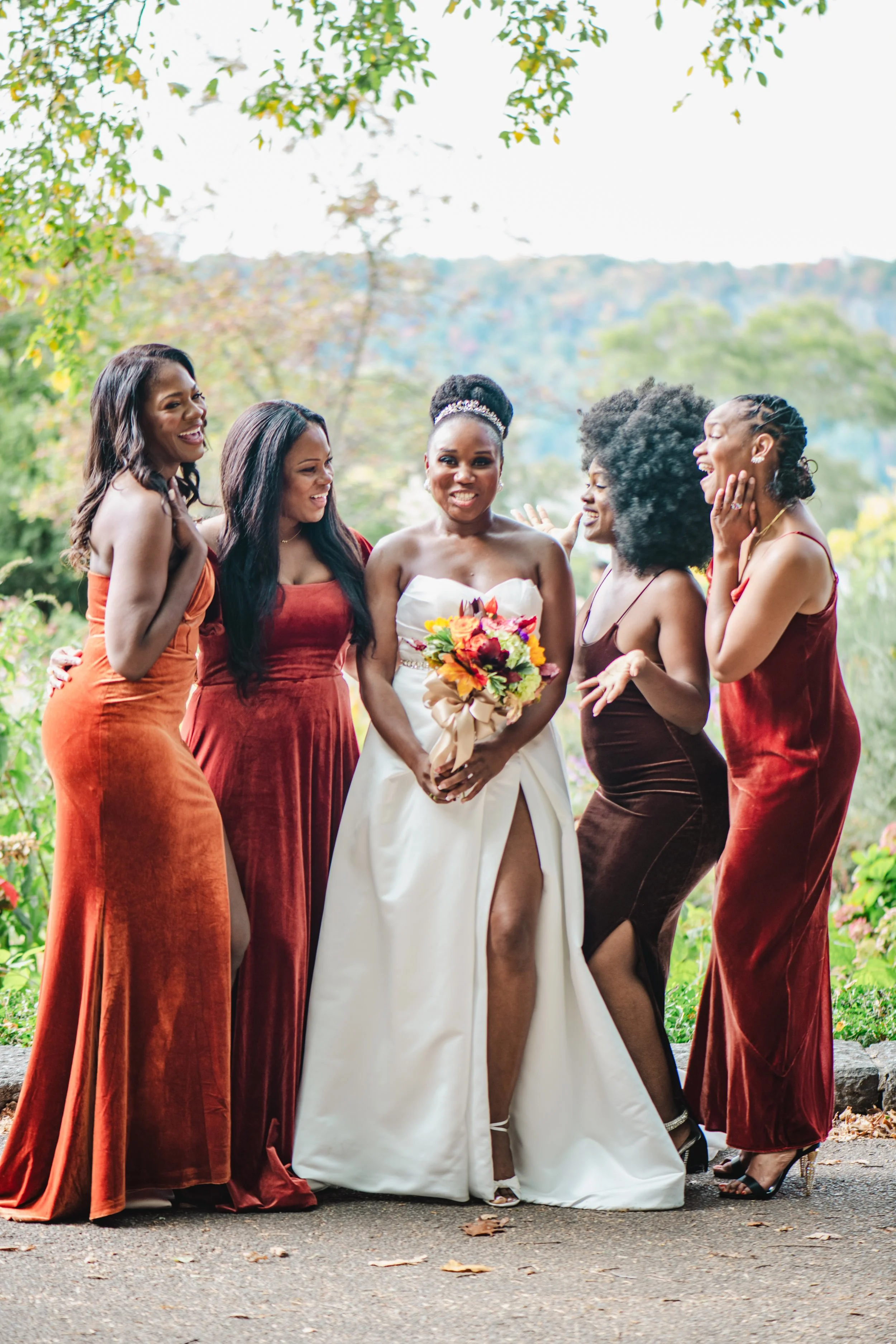 A bride in a white wedding gown holding a bouquet, surrounded by five women in various shades of red and brown dresses, outdoors with trees and hills in the background.