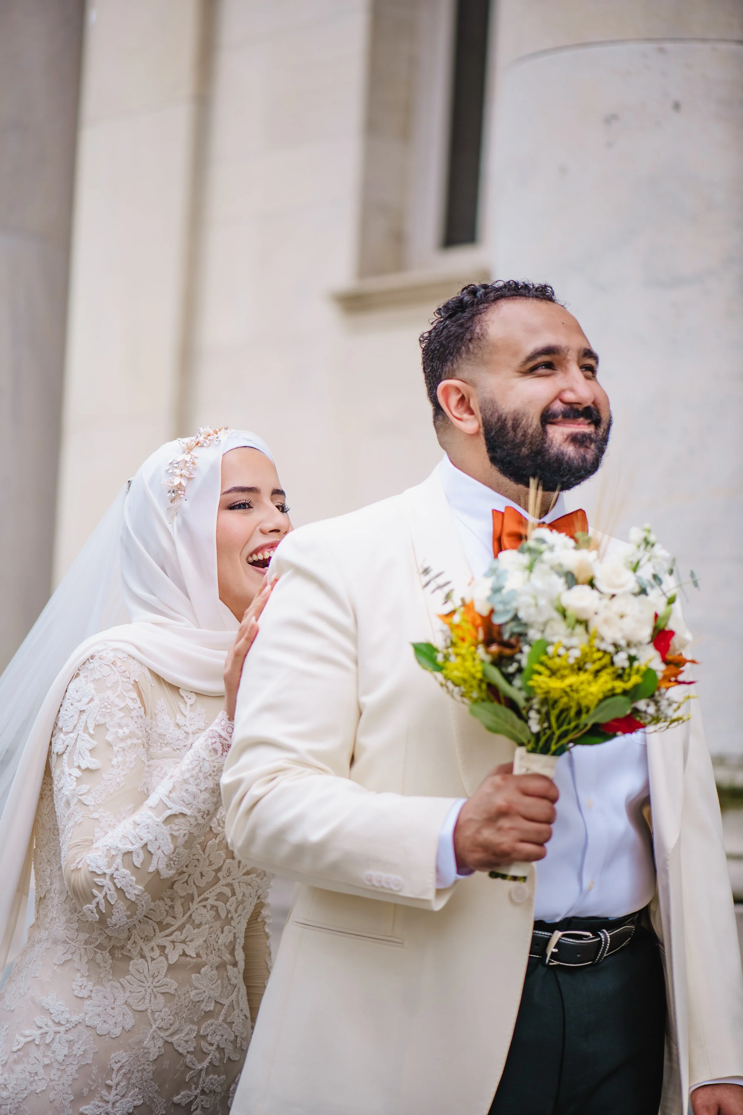 A newlywed couple embraces outdoors, with the man dressed in a dark suit and the woman in a lace wedding dress and floral crown, smiling affectionately at each other.