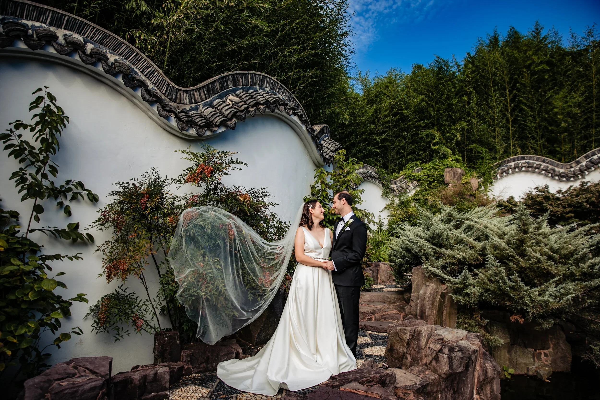 A bride and groom standing on rocky terrain, smiling and holding hands outdoors with lush greenery and a traditional Chinese-style wall behind them.
