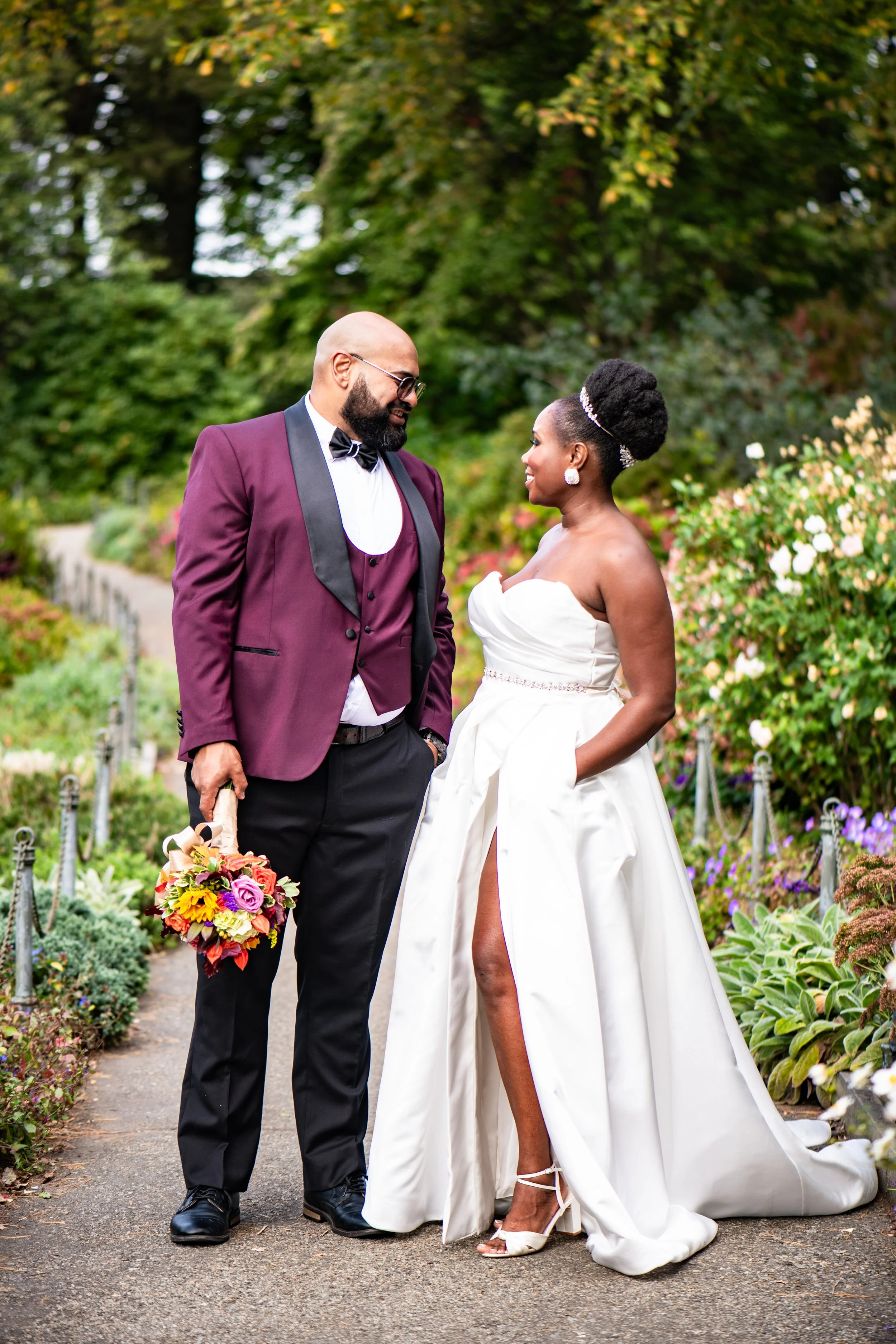 A bride and groom standing in a garden, gazing at each other. The bride is in a strapless white wedding gown with a high slit and high heels, and the groom is in a burgundy tuxedo jacket with black lapels, a vest, and trousers, holding a bouquet. The background features greenery and flowering bushes.