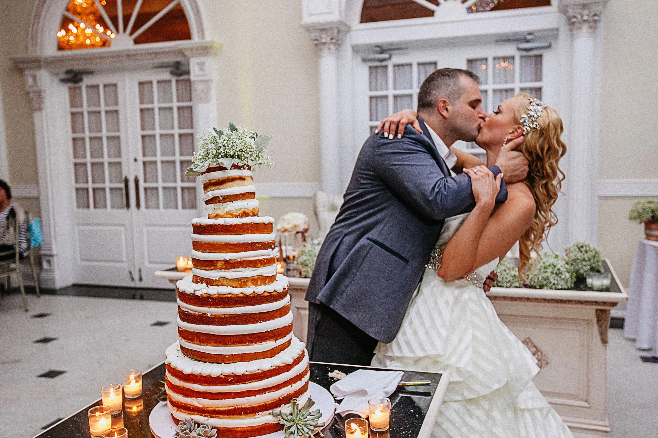 A bride and groom share a kiss during their wedding reception, with a tall naked wedding cake decorated with flowers and candles in the foreground.