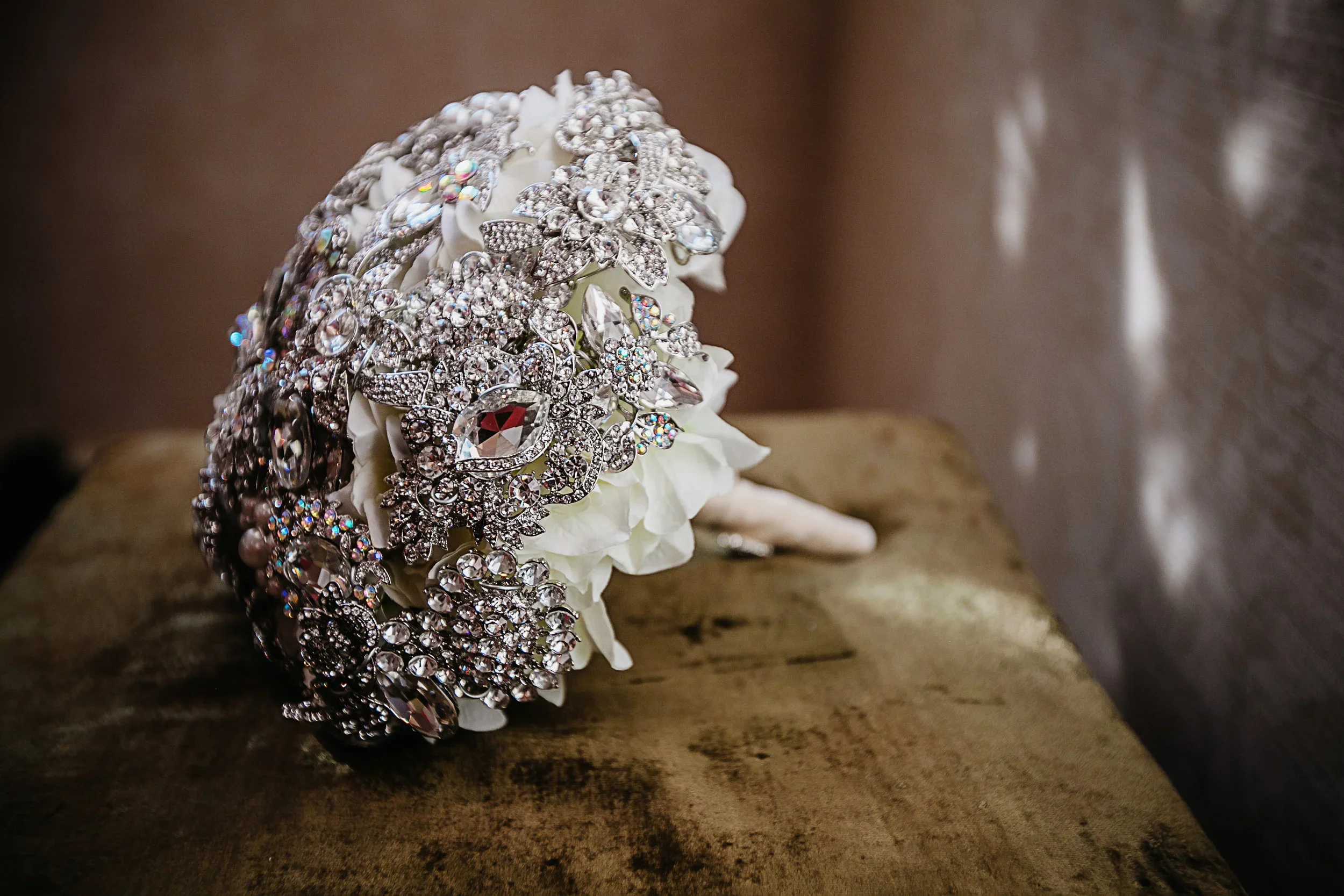 A bouquet of white flowers decorated with multiple layers of sparkling silver and rhinestone jewelry, resting on a wooden surface with a soft-focus background.