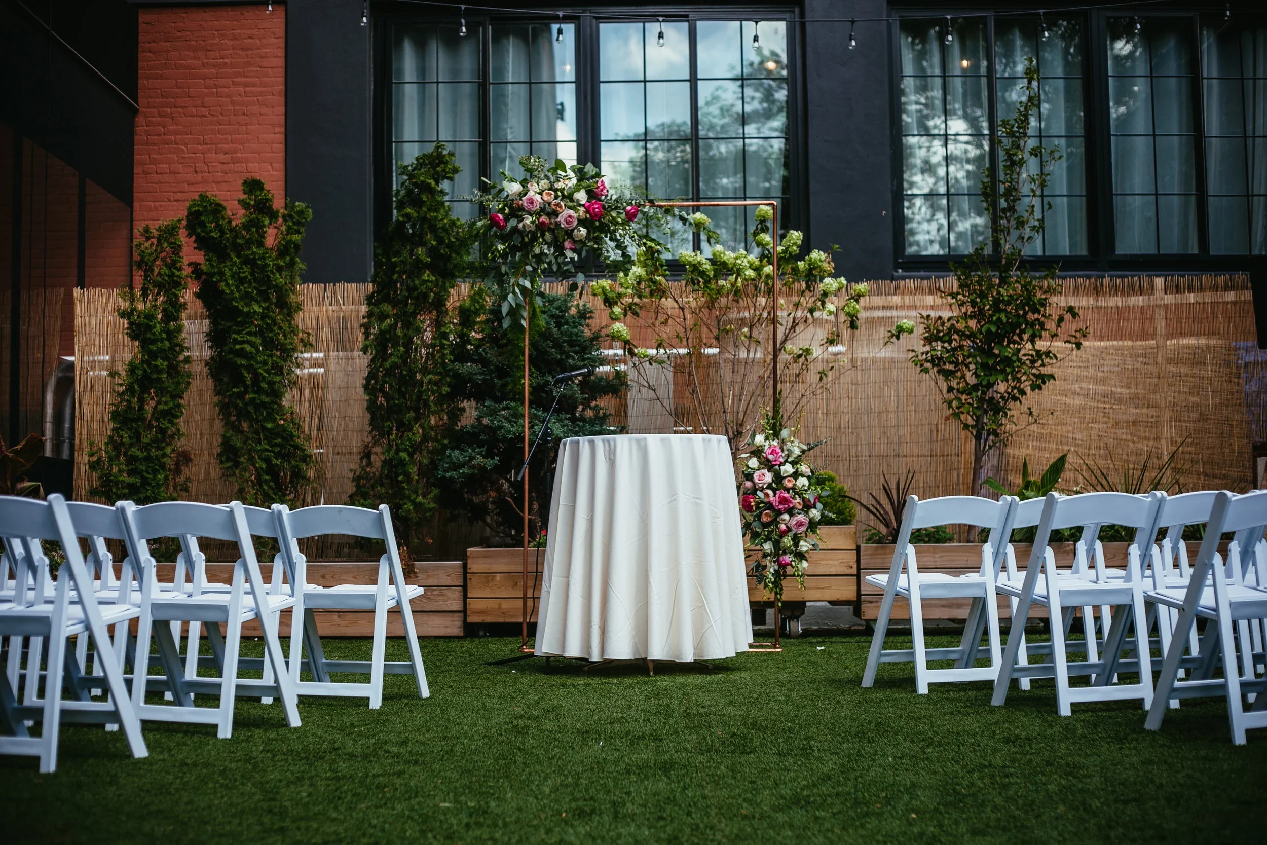 Empty outdoor wedding ceremony setup with white folding chairs arranged on green grass, a small round table with a white tablecloth, floral arrangements, and a backdrop with large windows and trees visible outside.