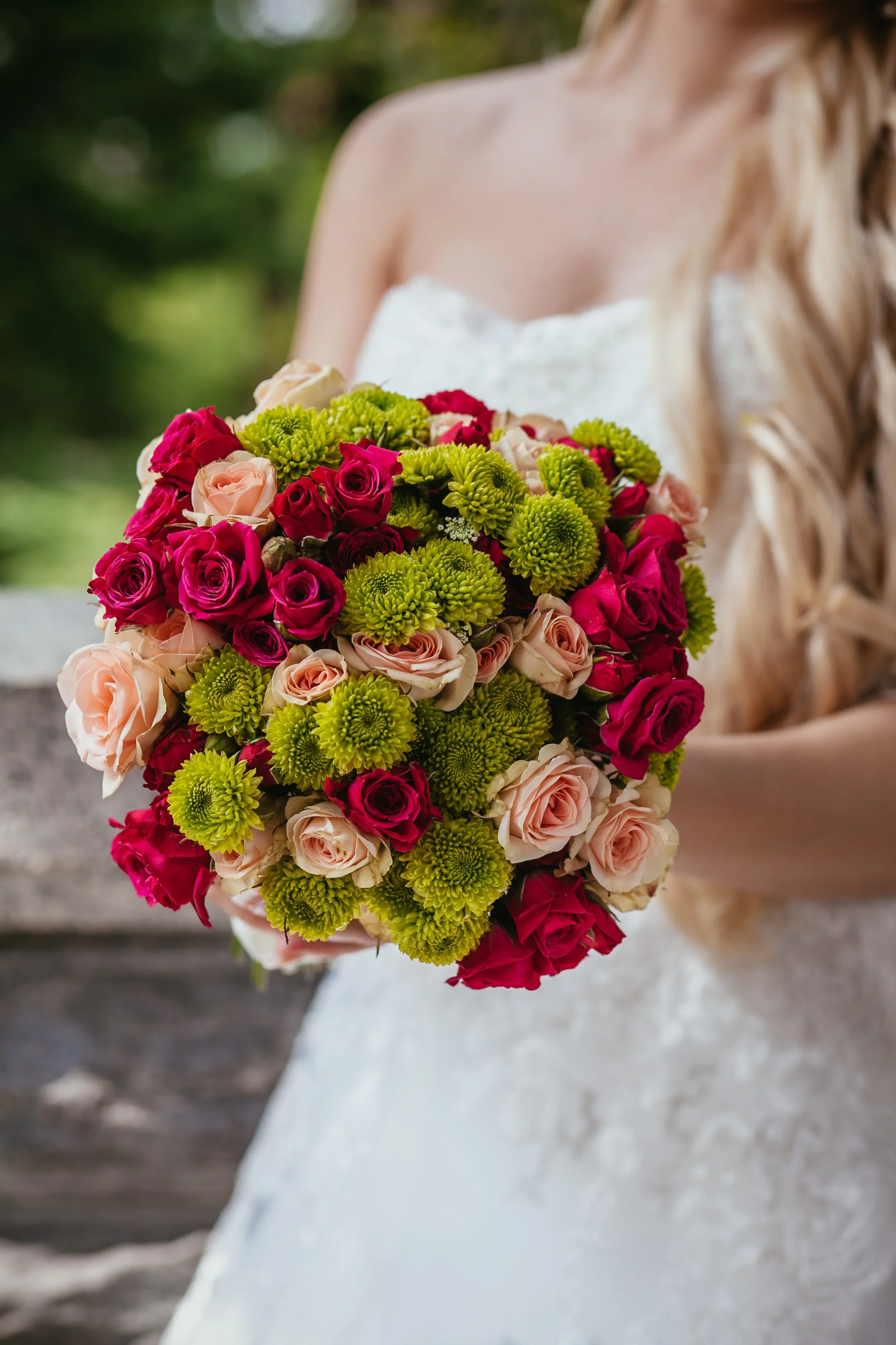 A long dining table set for a celebration outdoors on a dirt path, surrounded by greenery and string lights, with a rustic wooden shelter and tall trees in the background.