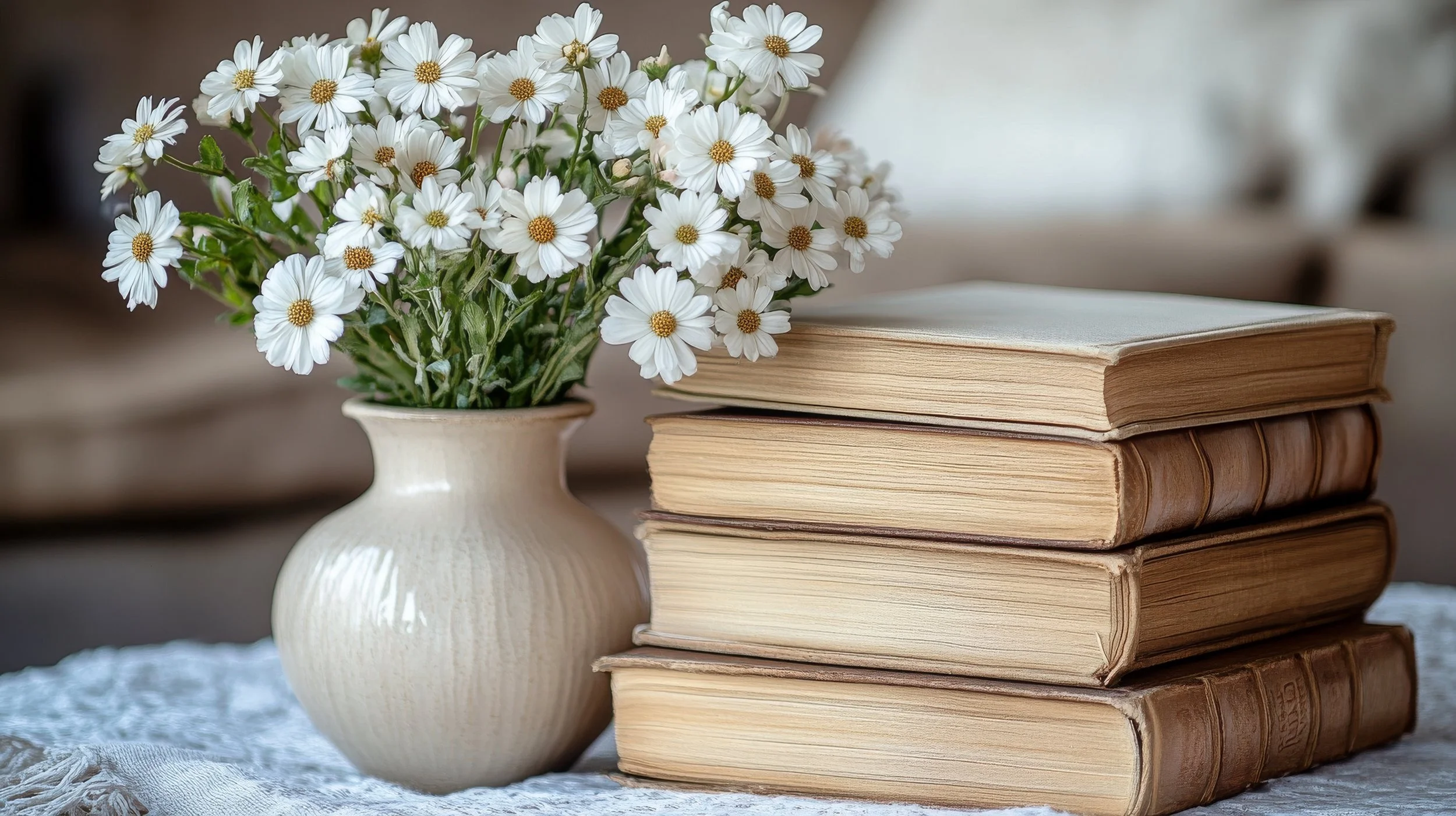 A beige vase with a textured surface holding a bouquet of white daisies, placed beside a stack of four old, hardcover books on a white lace tablecloth.
