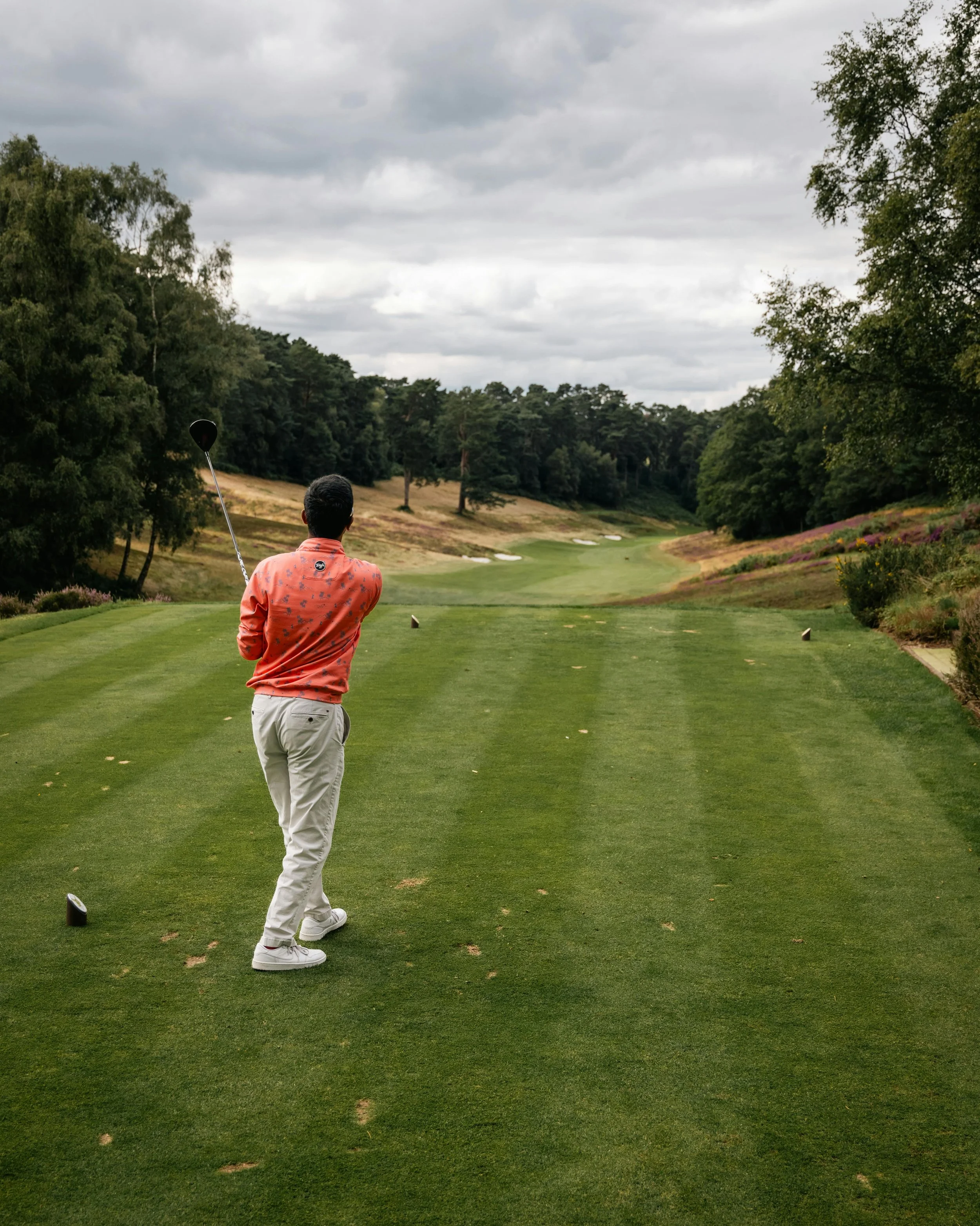 Man with short black hair wearing a salmon pink patterned shirt and white pants preparing to hit a golf ball on a lush green golf course, with trees and cloudy sky in the background.