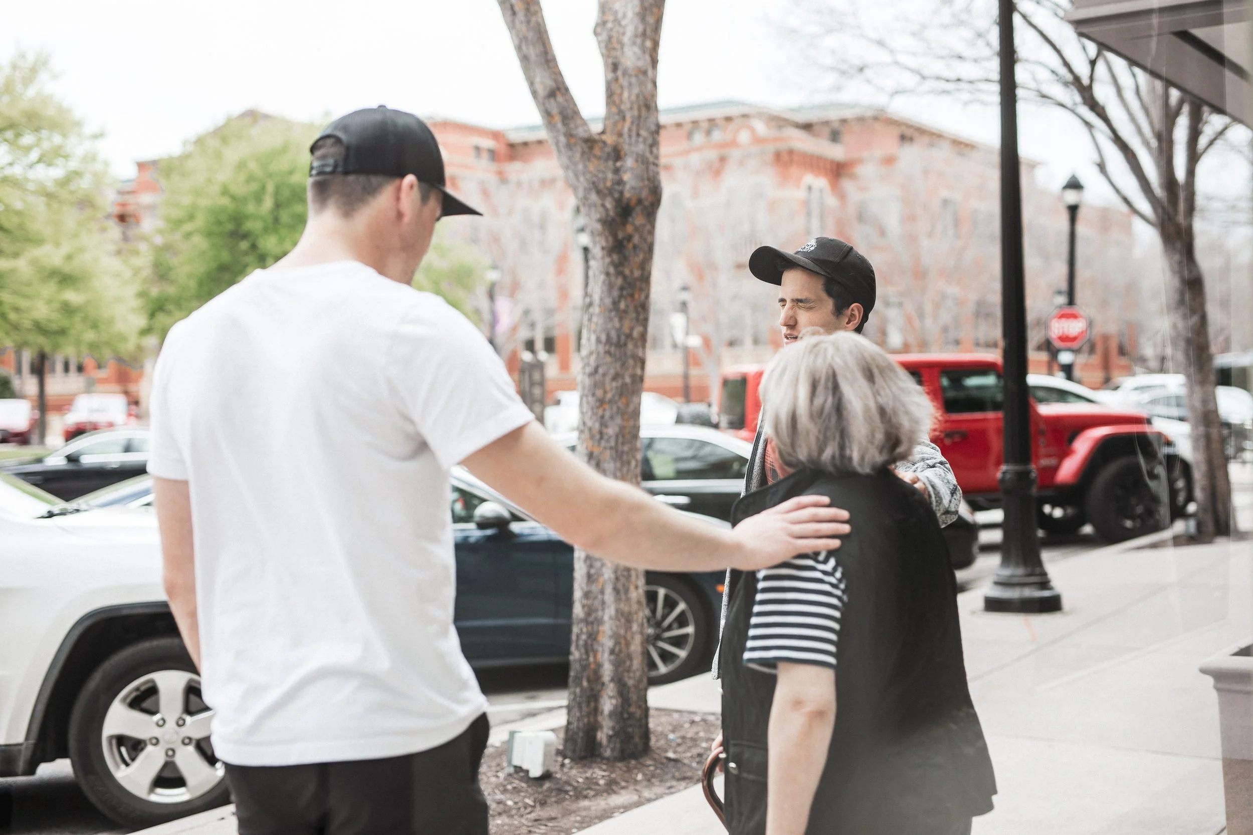 Three people, two young men and an older woman, are standing outdoors on a sidewalk, engaged in conversation. The young men are wearing caps and casual clothing, one in a white t-shirt and the other in a striped shirt with a dark vest. The woman has gray hair. There are cars parked along the street, trees, and a historic brick building in the background.