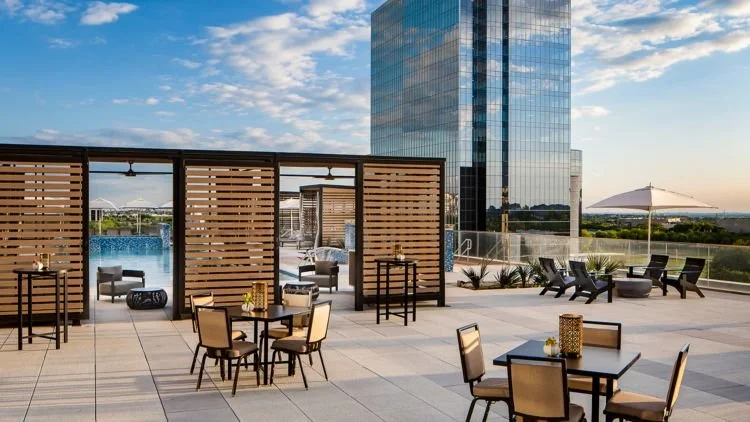 Rooftop terrace with tables and chairs, a pool area with privacy screens, a large modern glass building, and an umbrella with additional seating, under a partly cloudy sky.