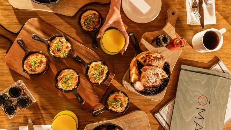 Top-down view of a wooden table set with various small skillet dishes, a glass of orange juice, a plate of bread and pastries, bottles of hot sauce, a mug of coffee, and a menu.