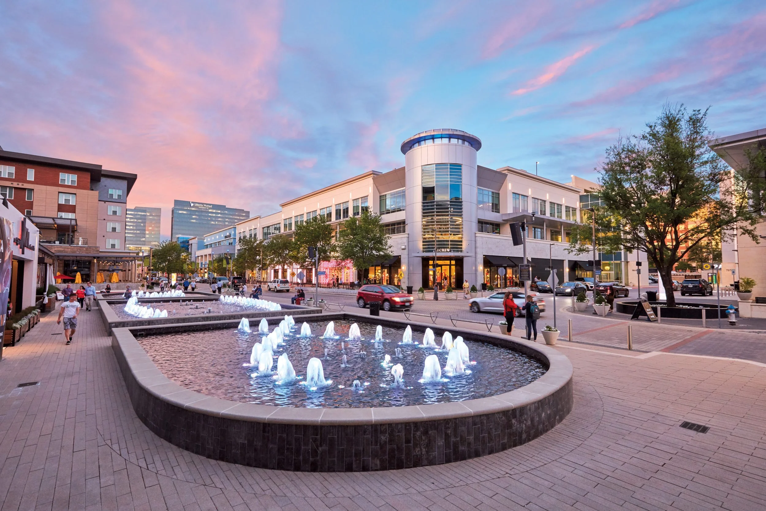 Exterior view of a luxury shopping mall with designer stores including Louis Vuitton and Gucci, with pedestrians and parked cars in front.