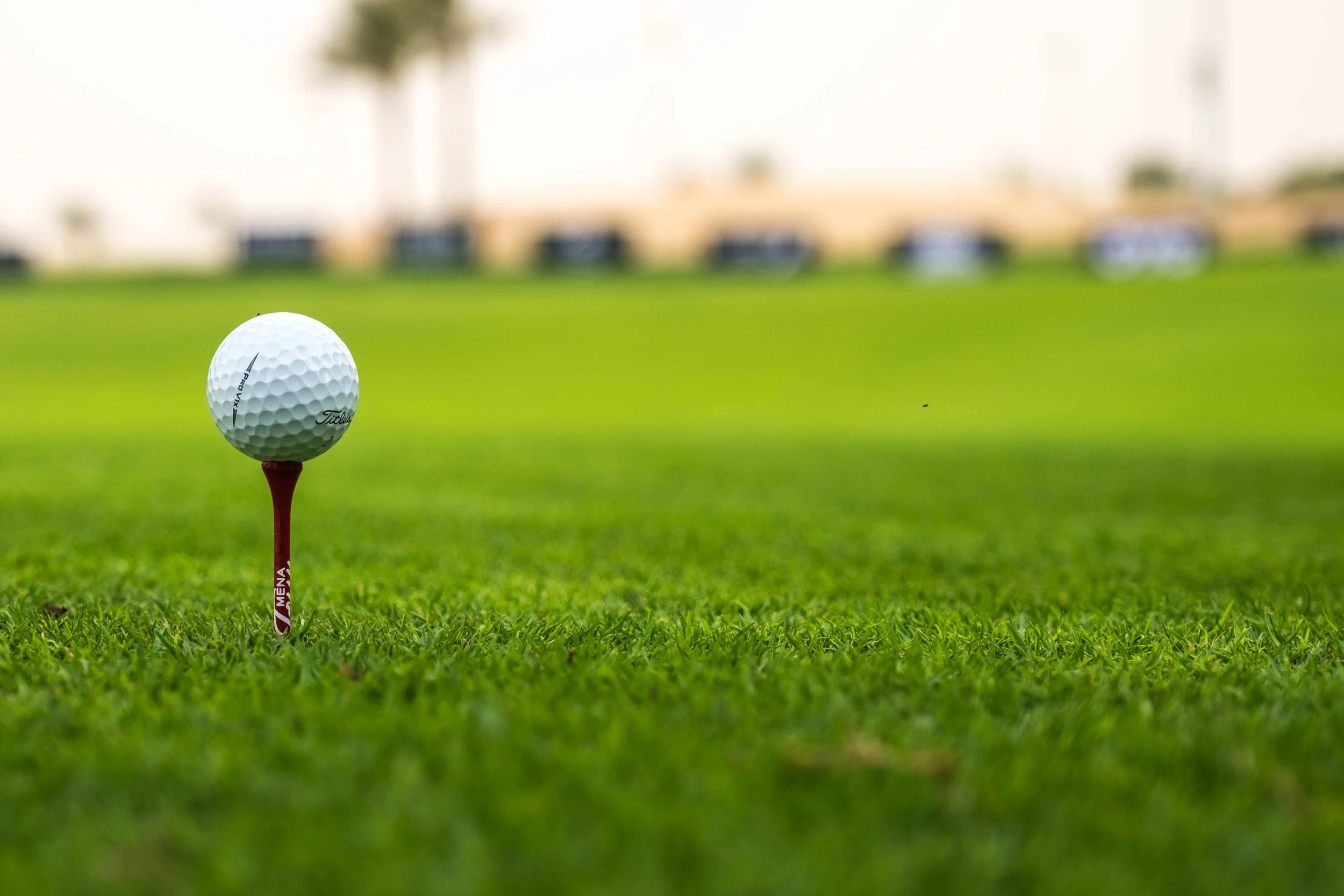 Man with short black hair wearing a salmon pink patterned shirt and white pants preparing to hit a golf ball on a lush green golf course, with trees and cloudy sky in the background.