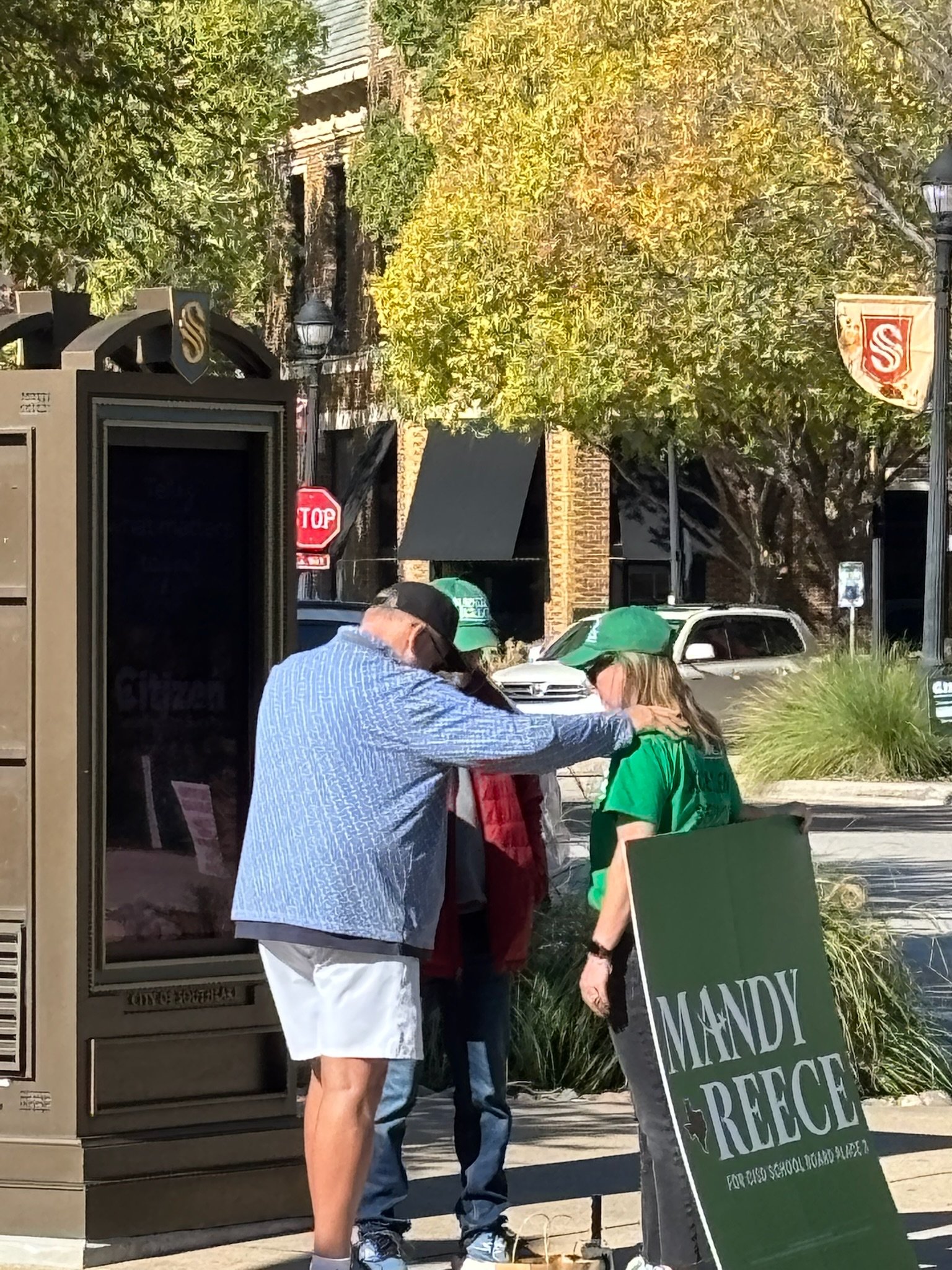 People waiting in line outside a building, with a person holding a green sign that reads 'MANDY'S PEACE' and a woman in a green shirt talking to a man in a blue shirt. There are trees, a stop sign, and parked cars in the background.