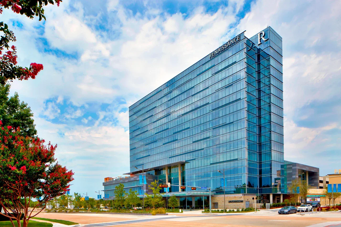Modern glass building with the word 'Renaissance' on top and front, reflecting the sky and surroundings, with trees and cars in the foreground.