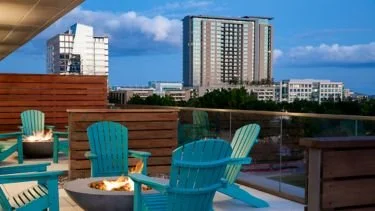 Rooftop outdoor lounge with wooden and teal Adirondack chairs, a fire pit, and city buildings in the background under a blue sky.