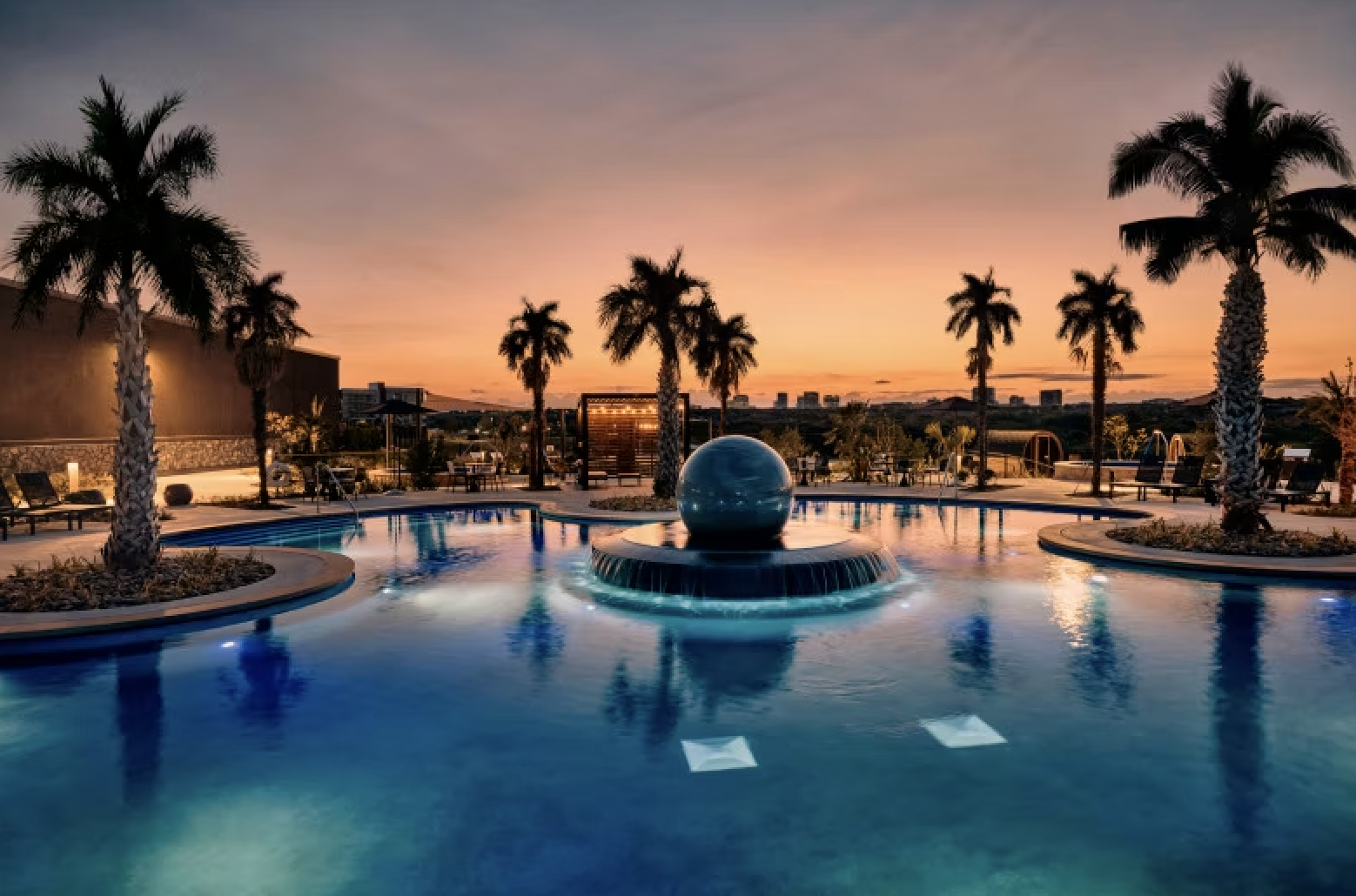 A swimming pool with a fountain and a large blue sphere, surrounded by palm trees during sunset, with lounge chairs and a city skyline in the background.