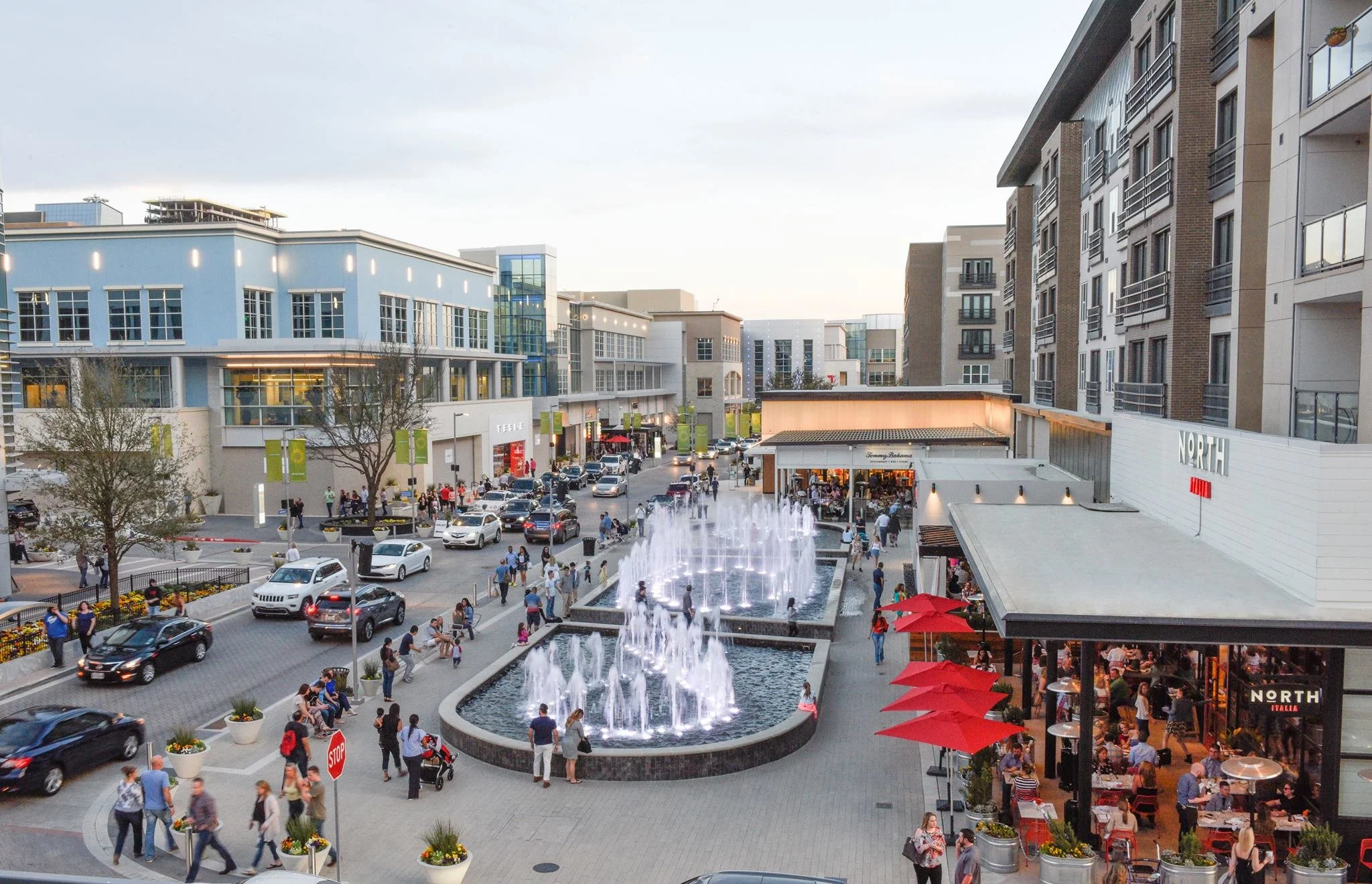 A busy shopping complex with a fountain in the foreground, and multi-story buildings with shops and restaurants on the sides, including a Nike store and an outdoor seating area with red umbrellas, populated with pedestrians and cars.