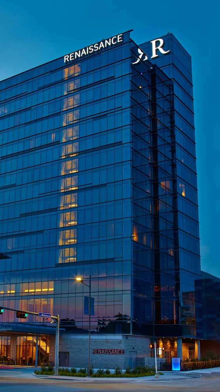 A modern glass office building with the company name 'Renaissance' on the top and a separate sign at the bottom, illuminated in the evening. The building is reflected on the glass facade, and the street in front has streetlights and traffic signals.
