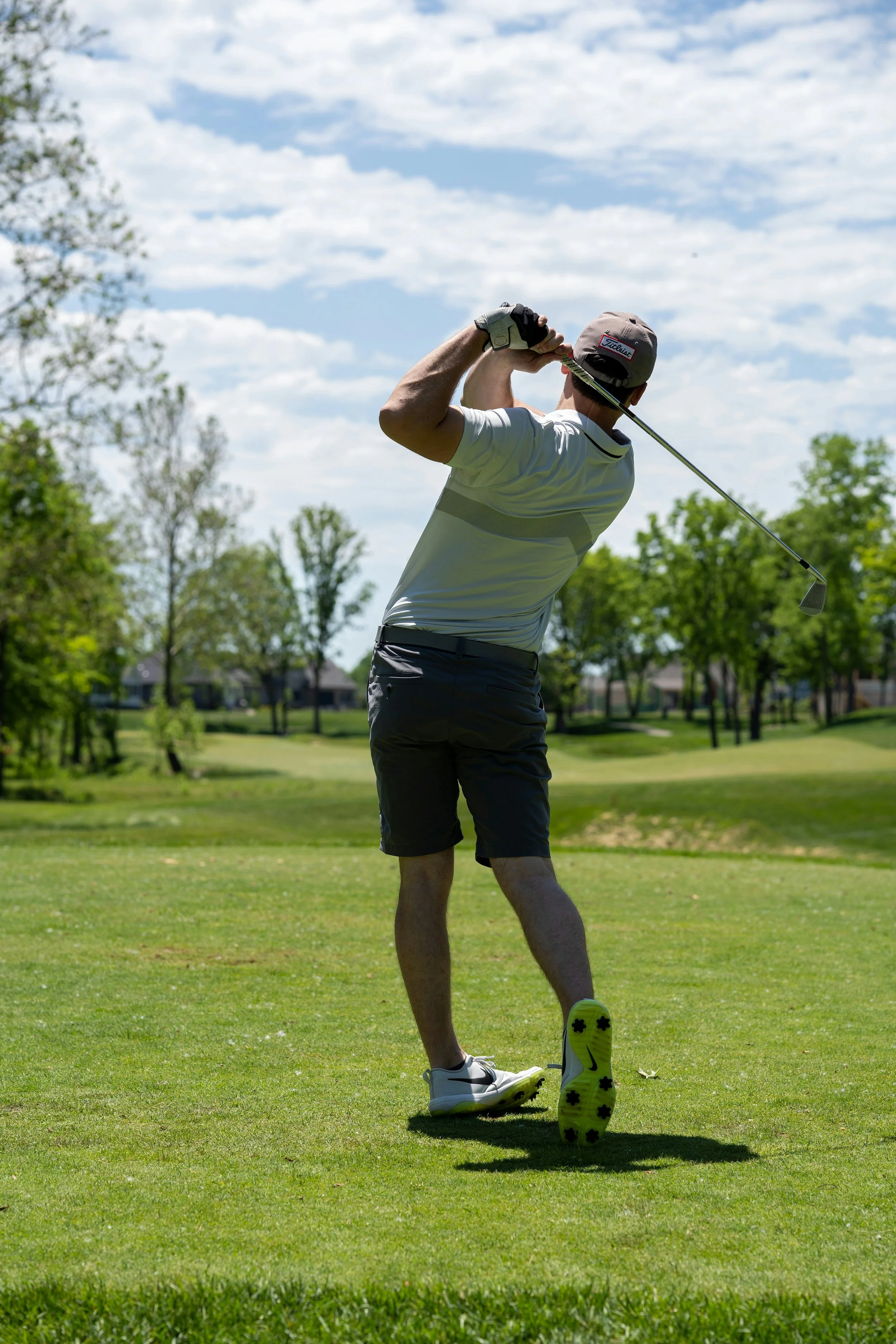 Male golfer in white shirt and gray shorts swinging a golf club on a sunny day with blue sky and scattered clouds, green trees, and well-maintained golf course in the background.