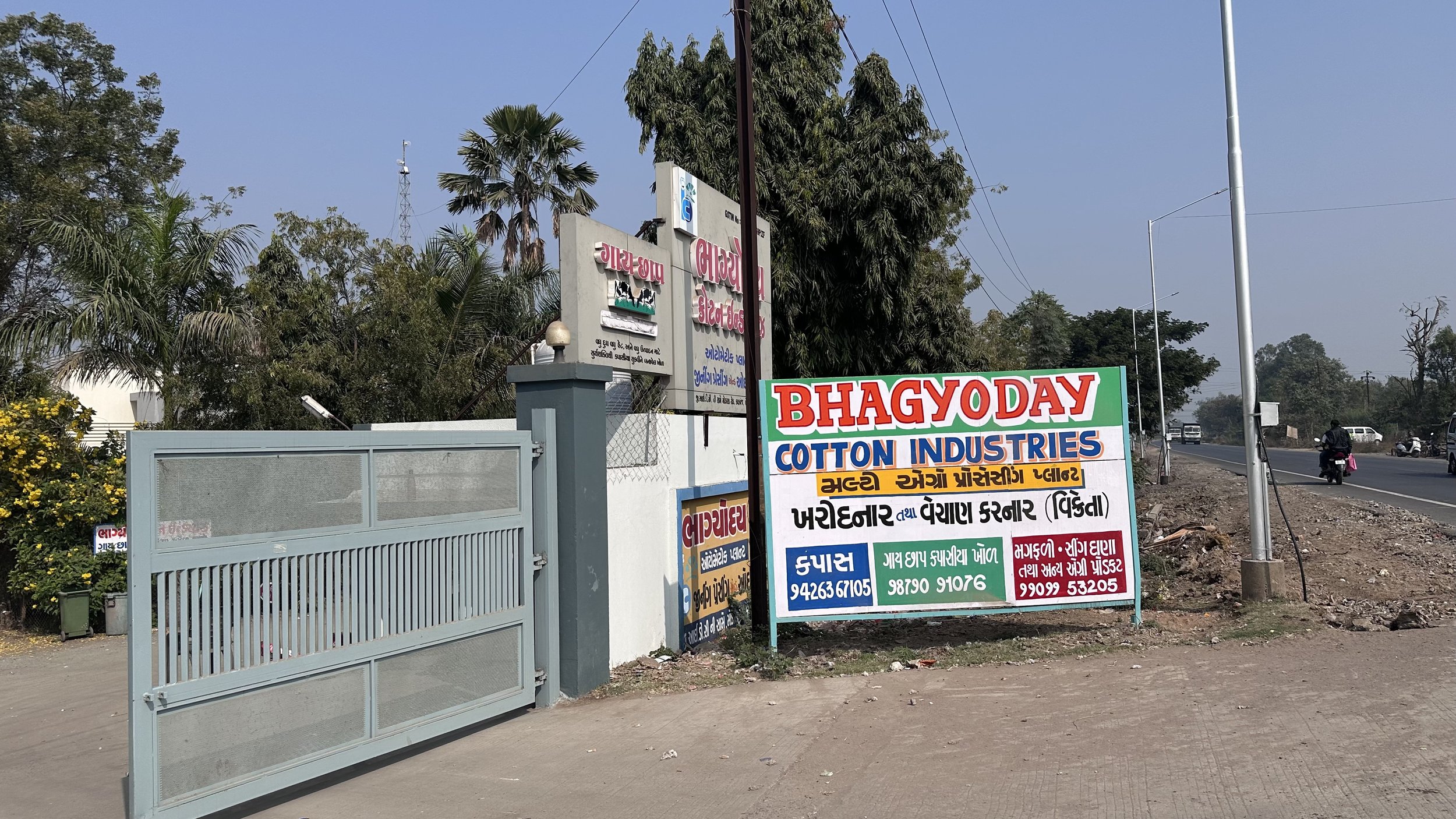 Signboard for Bhagyoday Cotton Industries advertising various services, with trees and a road in the background.