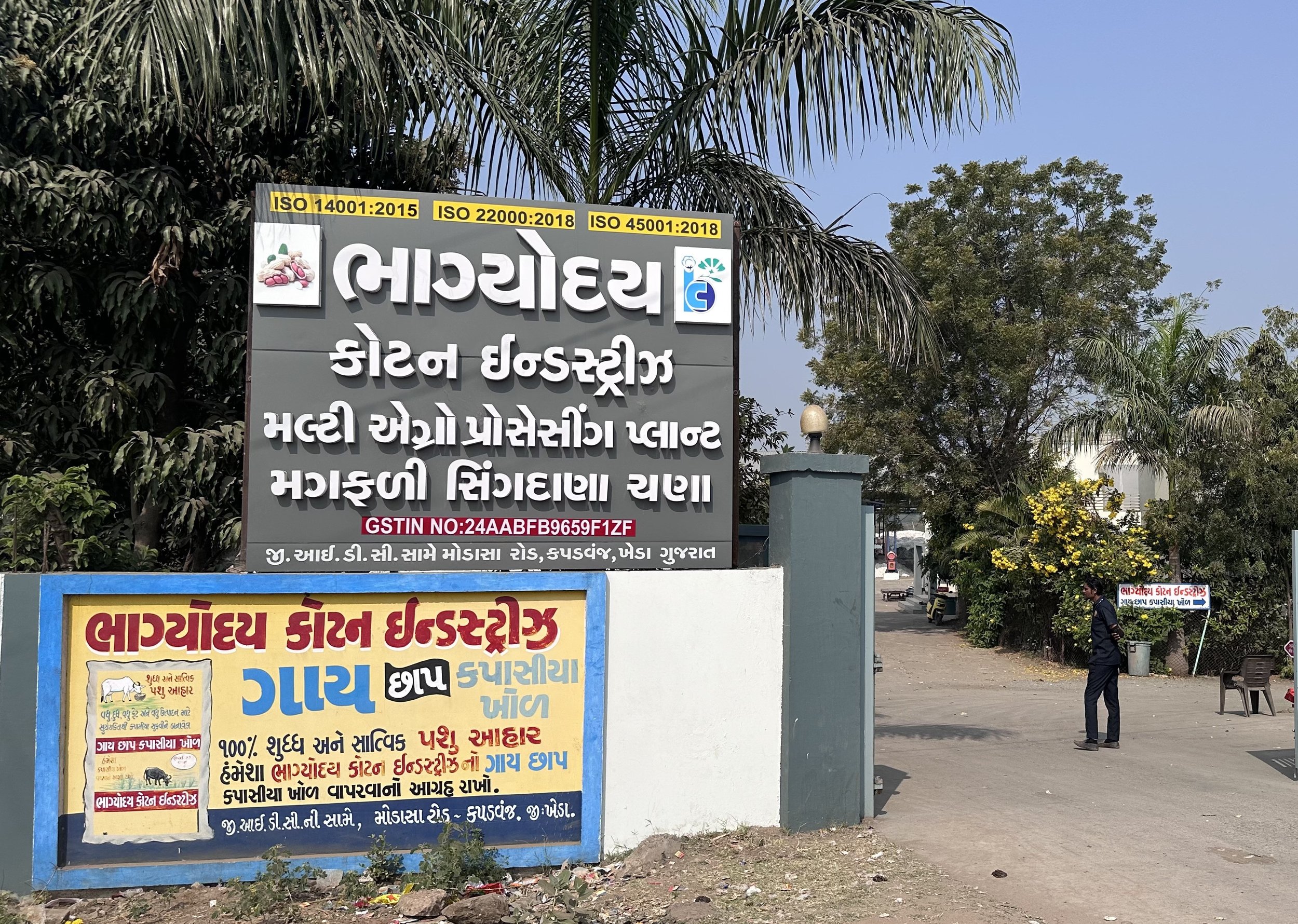 Signboard and poster in Gujarati language outside a building with trees in background, a man standing on the pathway.