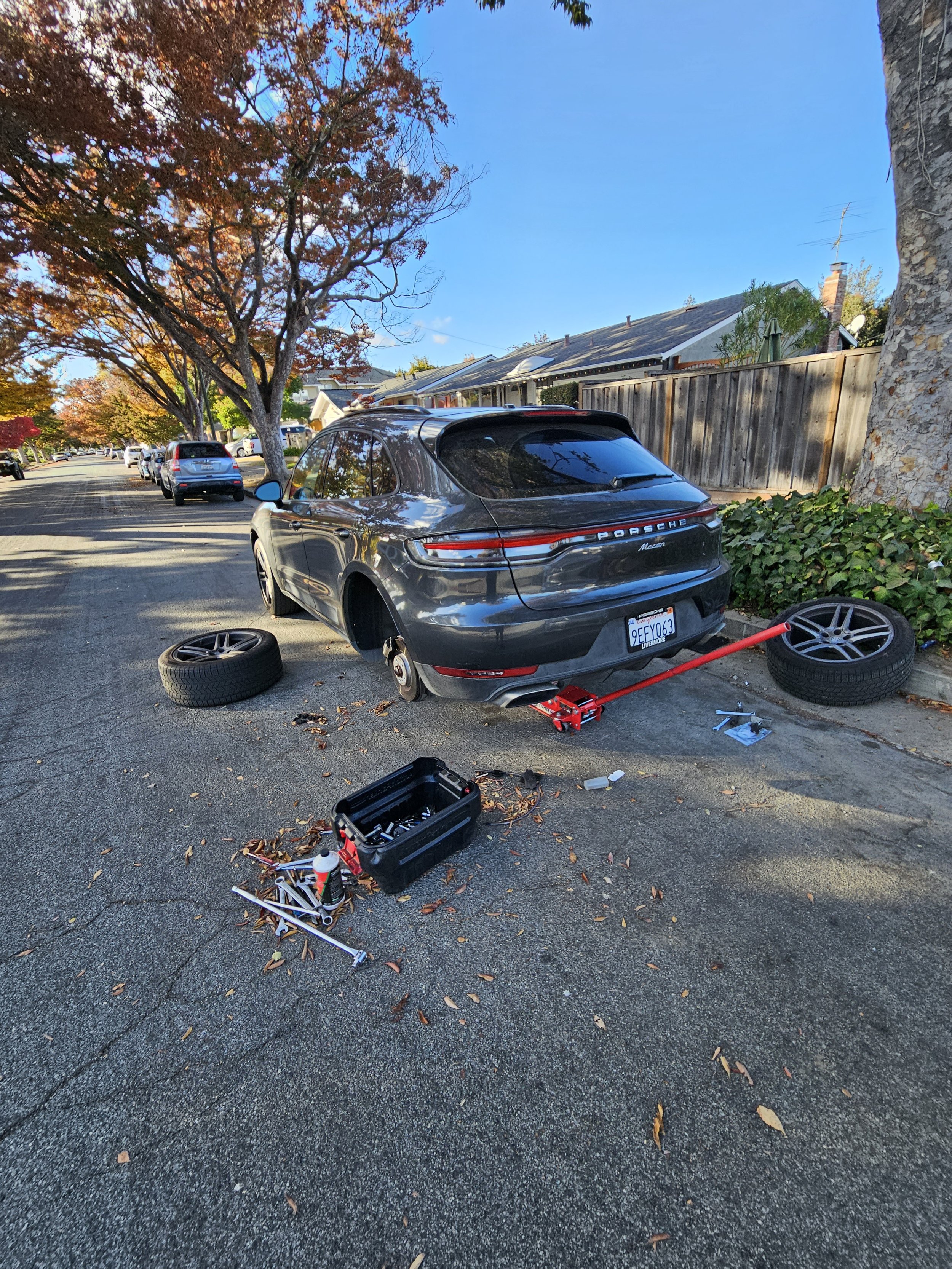 A black Porsche Macan SUV on a residential street with both rear wheels removed. The car is jacked up, with a red hydraulic floor jack beneath the rear. Tools and spare wheels are scattered around, including a toolbox and tire-changing equipment. The street has autumn trees and houses with fences.