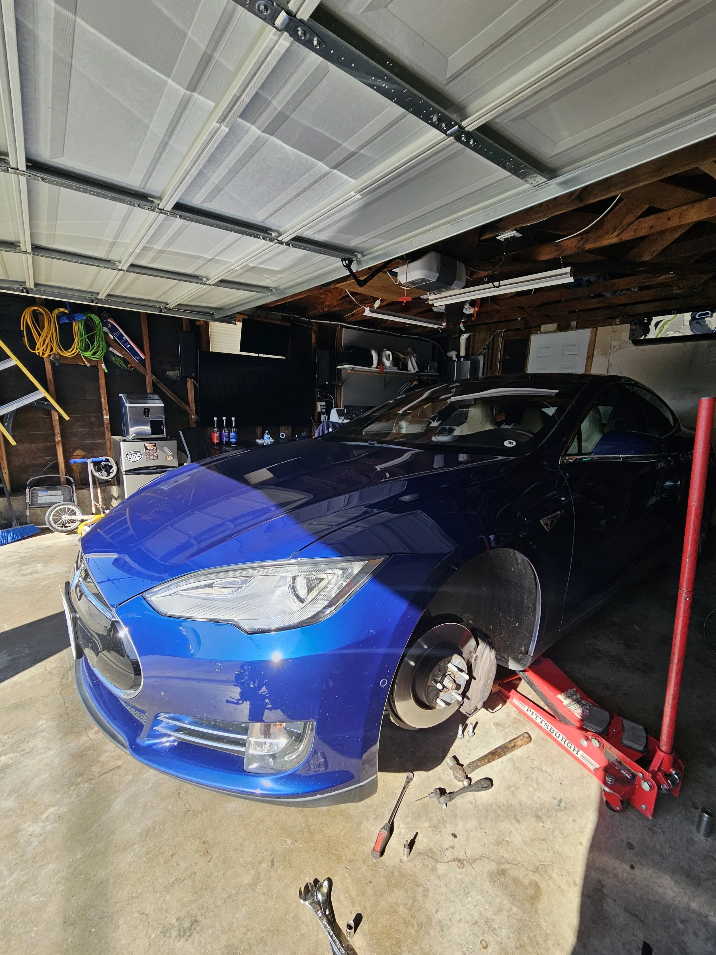 A blue Tesla car lifted on a red hydraulic car jack in a garage, with the front wheel removed. Tools are on the ground nearby, and the garage has equipment, shelves, and a garage door overhead.