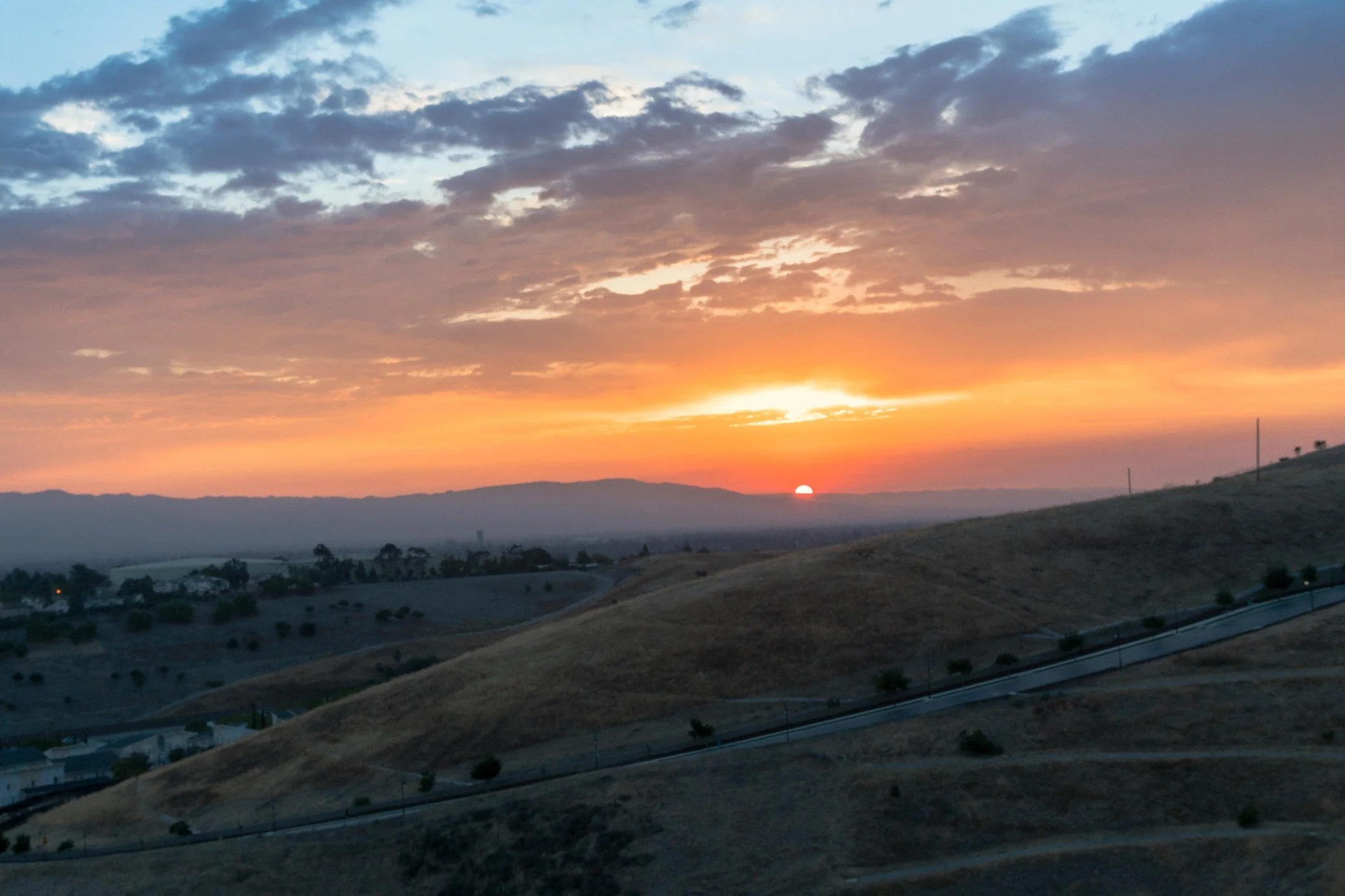 Sunset over rolling hills with scattered trees and a partly cloudy sky.