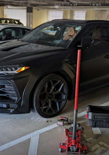 Black sedan parked in an indoor parking lot with a red hydraulic floor jack underneath it, a red service pole standing beside, and tools and equipment nearby.