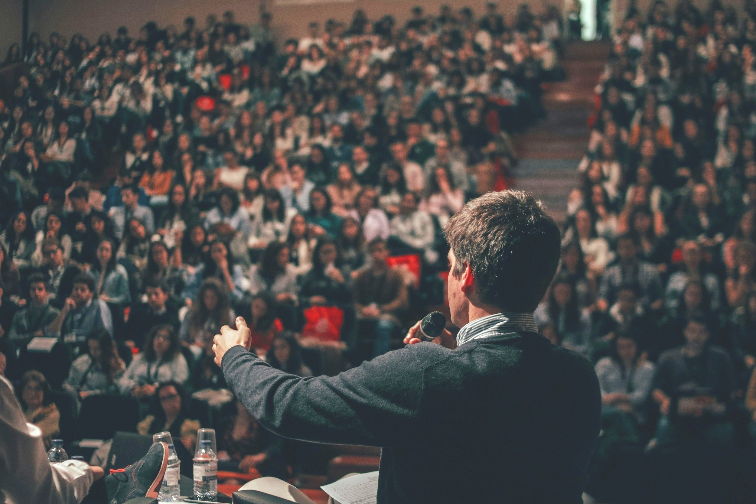 A man speaking into a microphone during a startup presentation or investor conference speech in a large auditorium filled with an audience.
