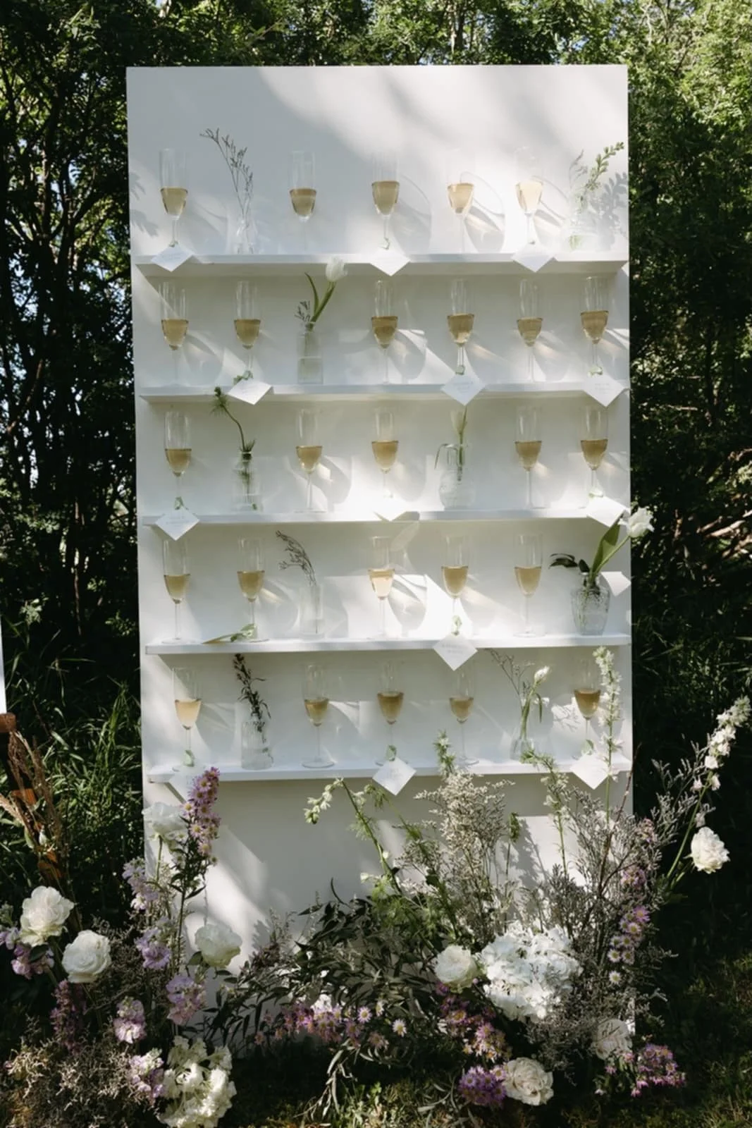 A white display board with multiple glasses filled with champagne, decorated with individual flowers and greenery in small vases, in an outdoor setting surrounded by trees and flowers.