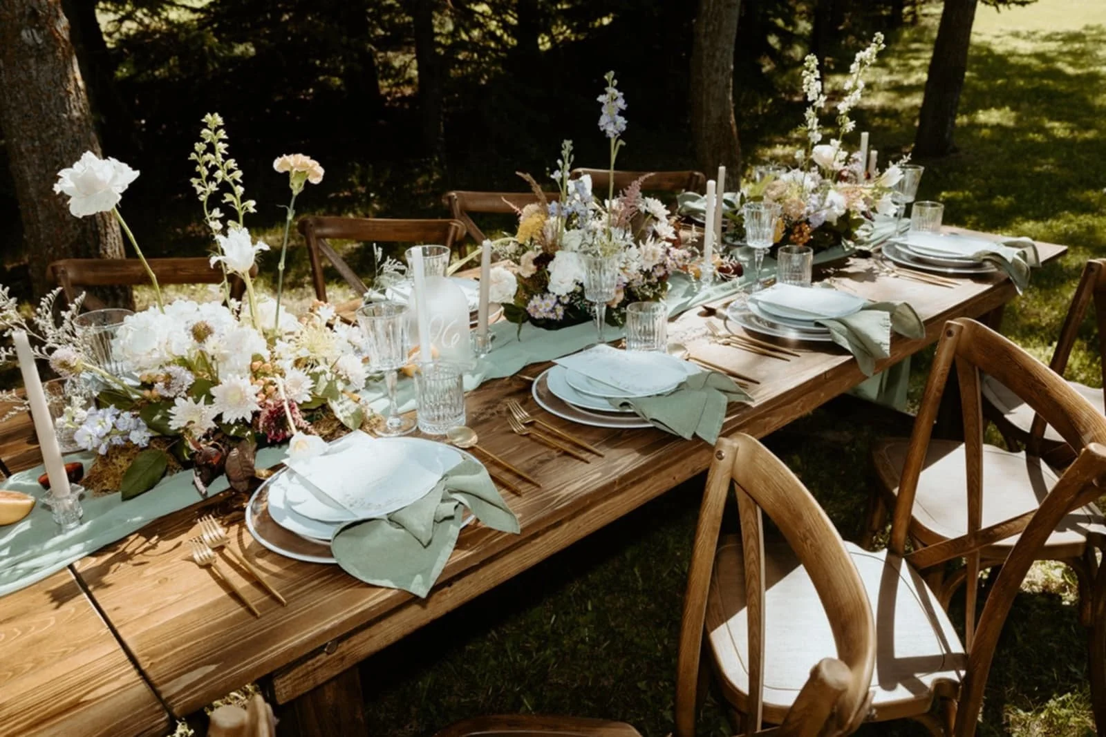 A long outdoor wooden table set for a meal, decorated with white and pastel floral arrangements, candles, and pastel napkins, surrounded by wooden chairs, in a forest setting.