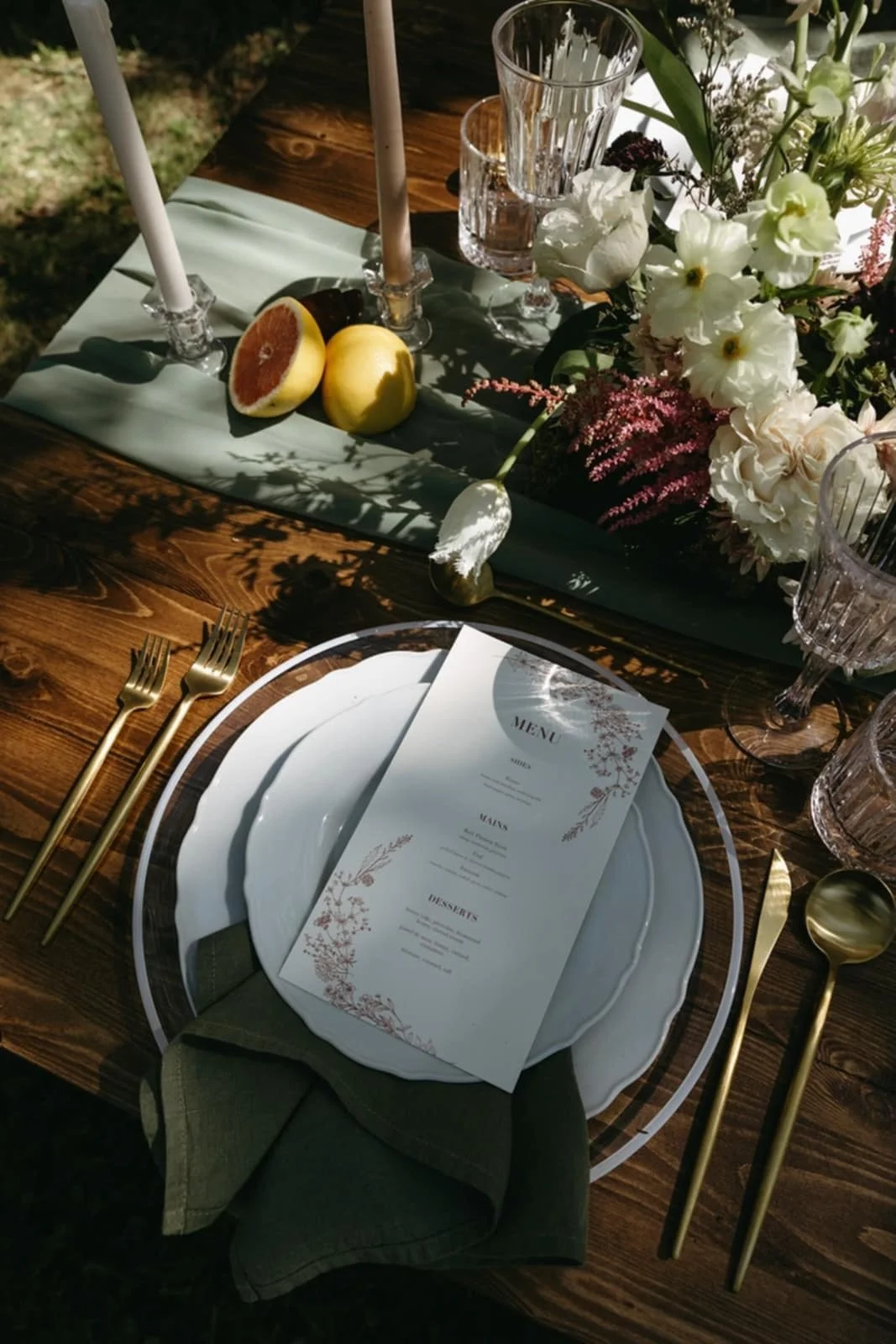 A beautifully set outdoor dining table with white plates, gold utensils, a menu, glassware, candles, and a floral centerpiece, with a cloth runner and fresh fruit including a lemon and a halved grapefruit.