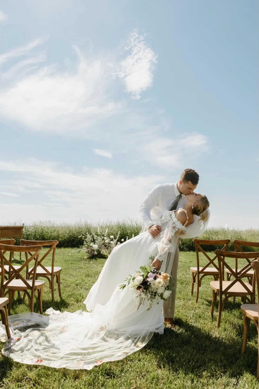 A bride and groom kissing outdoors at their wedding, with the bride holding a bouquet of flowers and the groom dressed in a white shirt and tie, surrounded by wooden chairs on green grass under a blue sky with clouds.