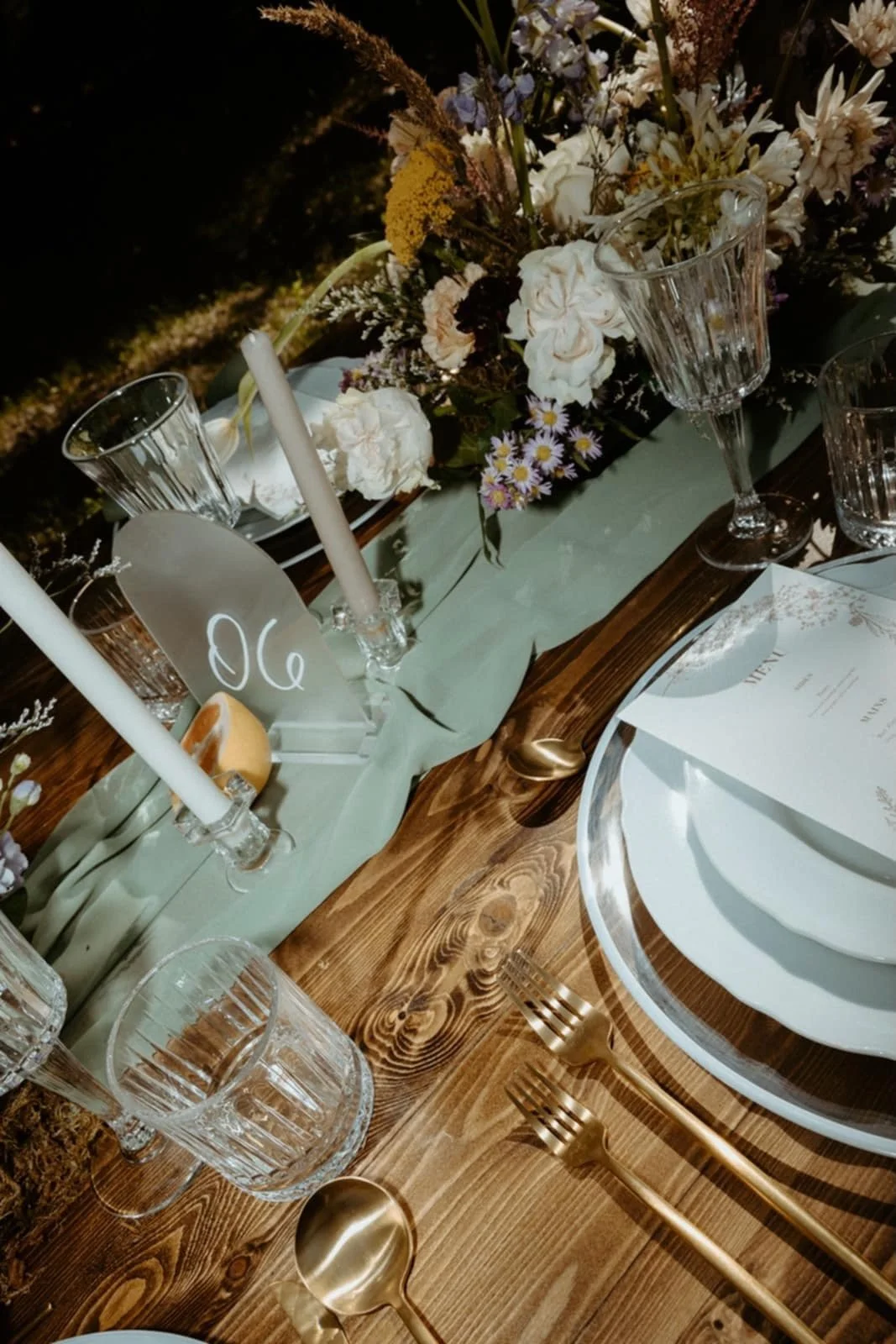 Table set for a formal event with white plates, gold cutlery, clear glassware, candlesticks, and a floral centerpiece with white, purple, and yellow flowers.