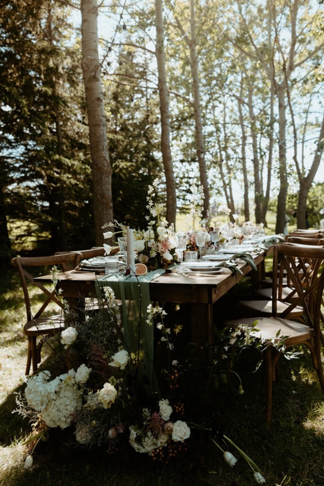 A rustic outdoor wedding table decorated with white and blush flowers, greenery, candles, and tableware, set in a wooded area with sunlight filtering through trees.