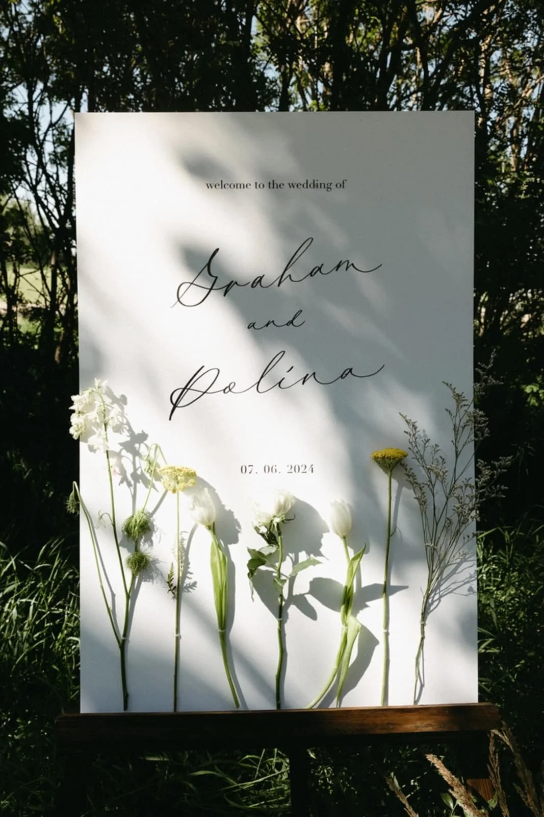 Wedding welcome sign with the names Graham and Palina, dated July 6, 2024, decorated with various white and yellow flowers against a backdrop of trees and sunlight.