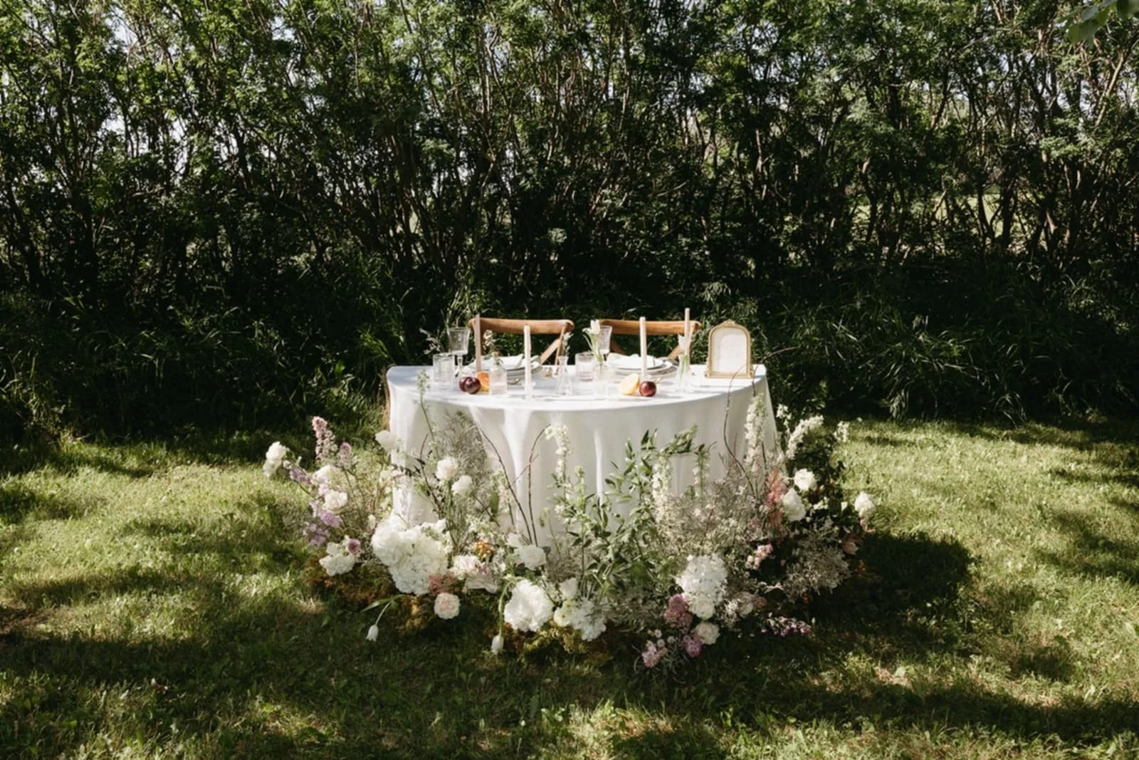 Round table with white tablecloth decorated with white and pink flowers, set with glasses, plates, and candles, placed outdoors on grass with bushes in the background.