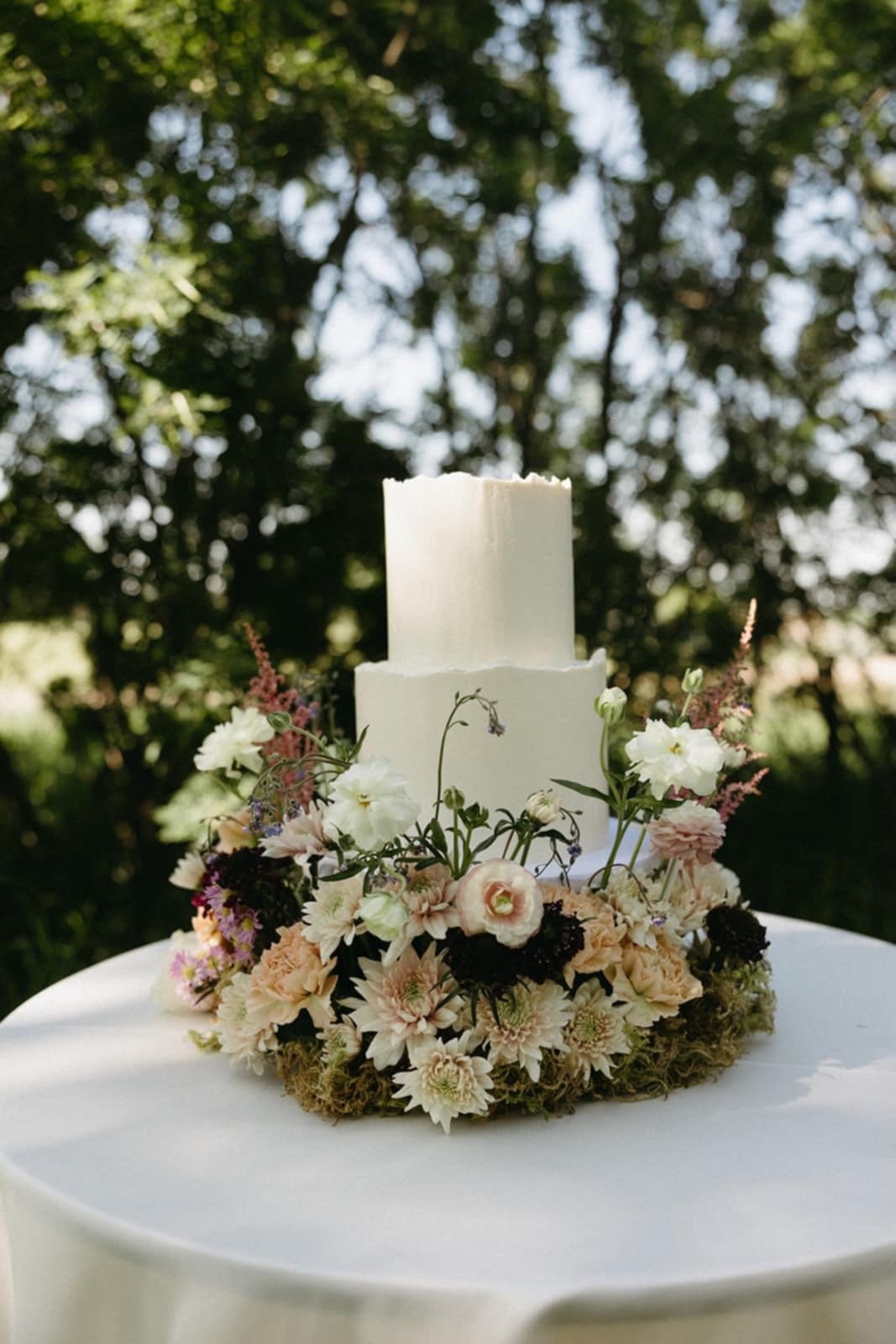 A white wedding cake with two tiers, decorated with various white, pink, and purple flowers, placed on a white table outdoors with a background of trees.