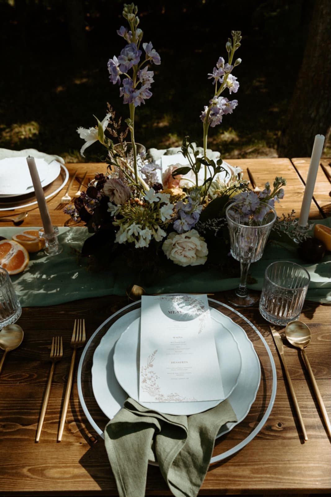 A table set outdoors with a centerpiece of purple, white, and pink flowers, gold cutlery, clear glassware, and a menu on a white plate with a beige napkin.