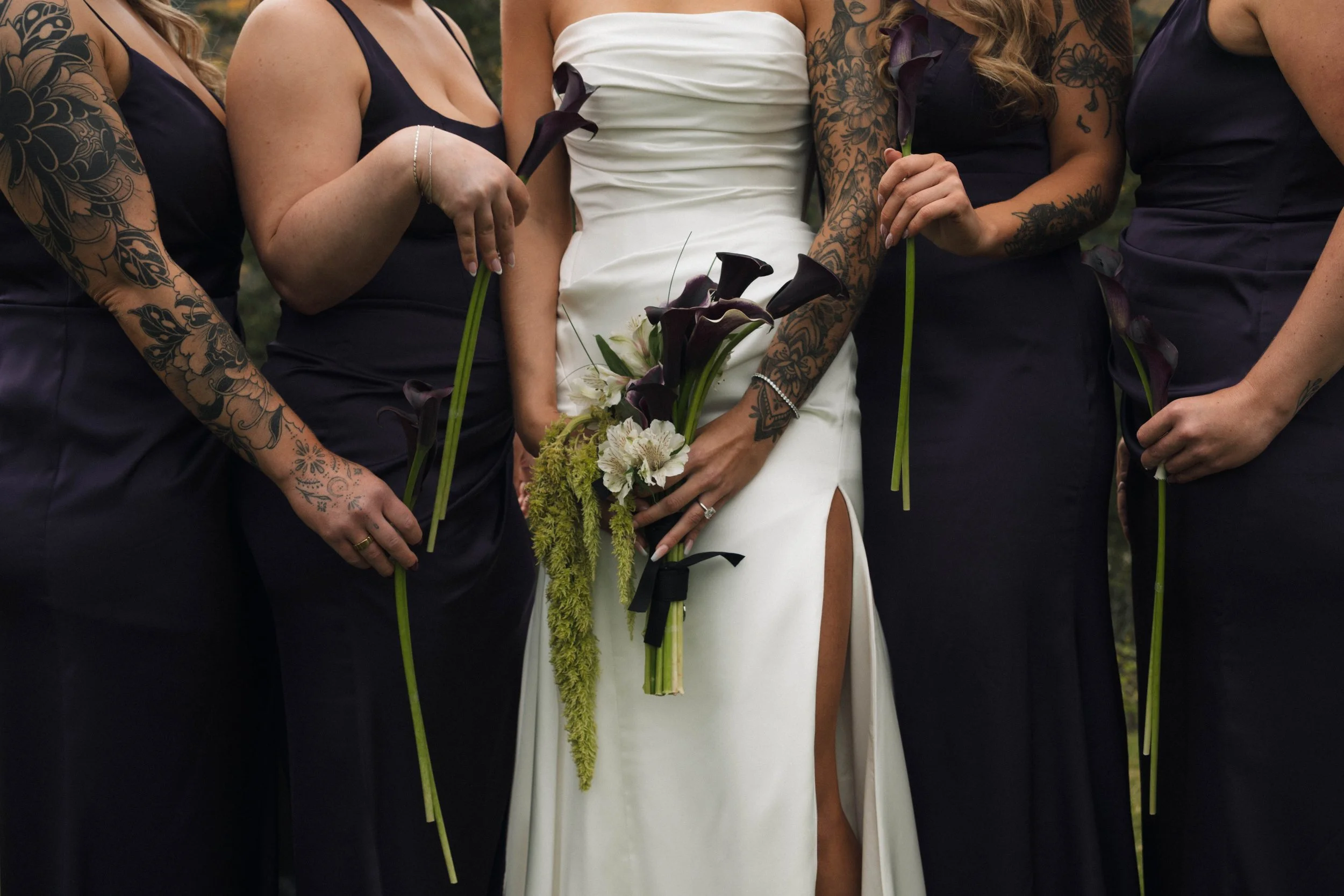 Close-up of a bride in a white dress surrounded by bridesmaids in navy dresses, holding dark purple calla lily flowers and a bouquet.