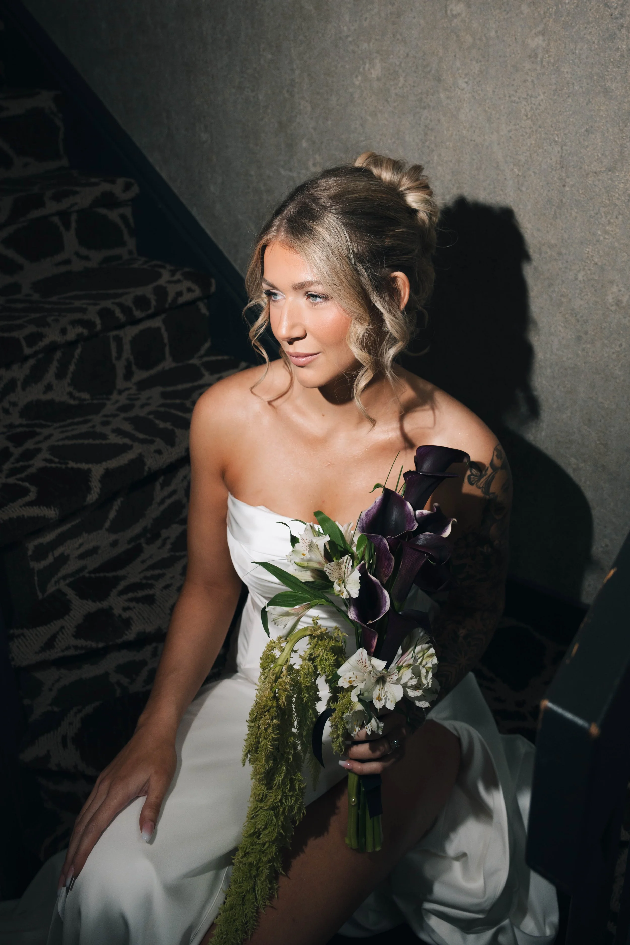 A woman in a strapless white dress sitting on a staircase holding a bouquet of dark purple calla lilies, white flowers, and greenery, with her blonde hair styled in loose curls.